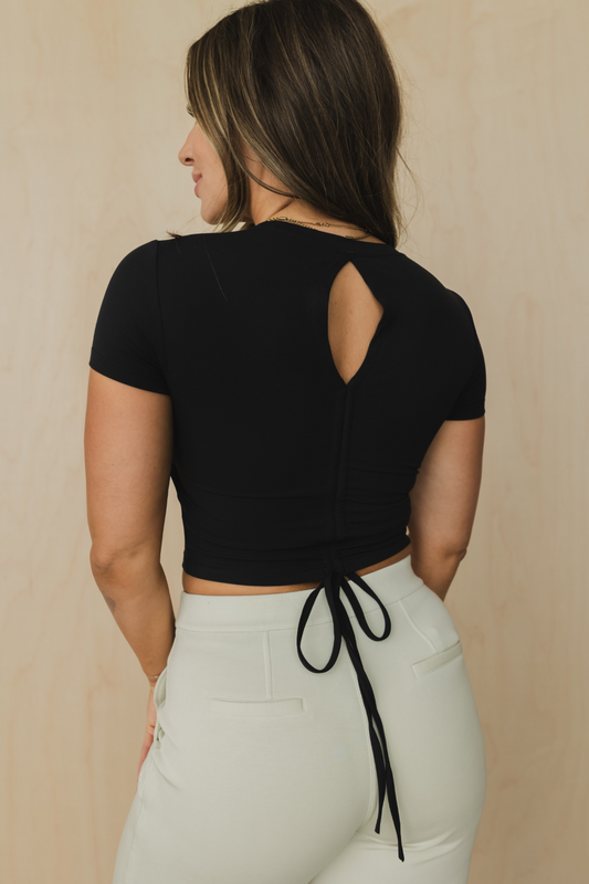 back view of a woman wearing a black crop top with cut-out design and light gray pants against a beige background