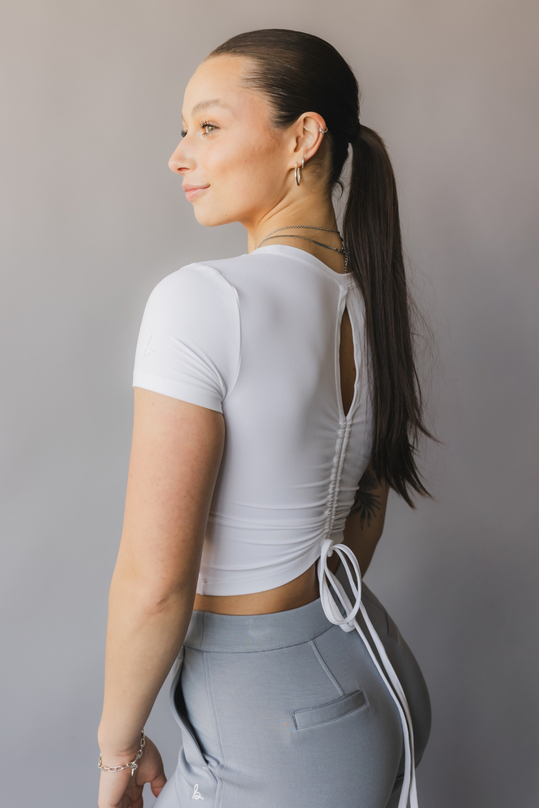 side view of a woman wearing a white crop top with cut-out design and light gray pants against a beige background