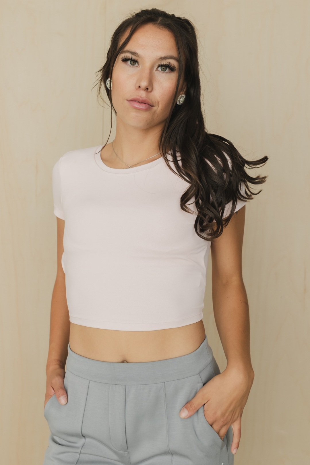 front view of a woman wearing a light pink crop top and gray pants against a beige background