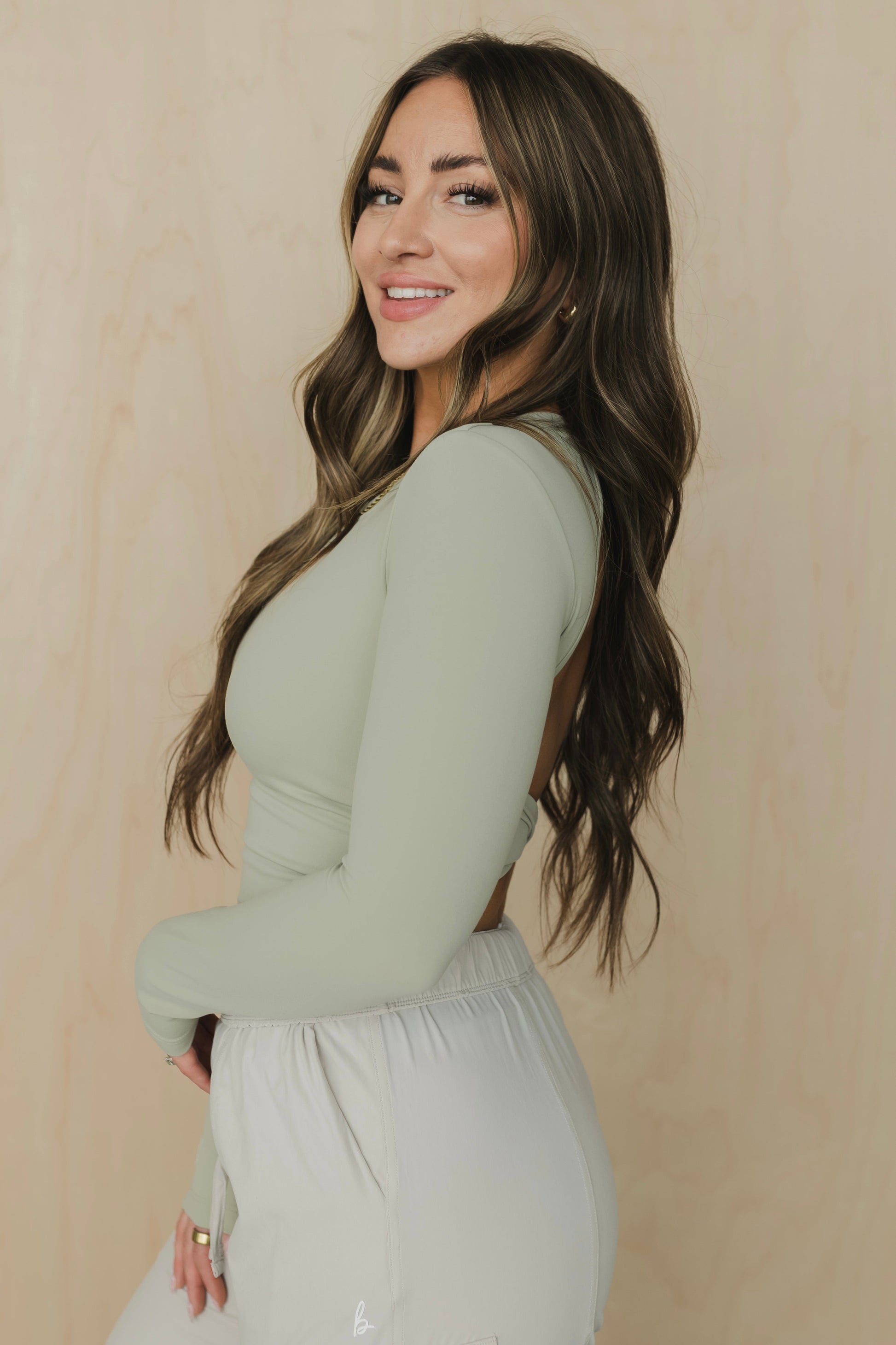 side view of a woman wearing a light green long-sleeve top and white pants against a beige background