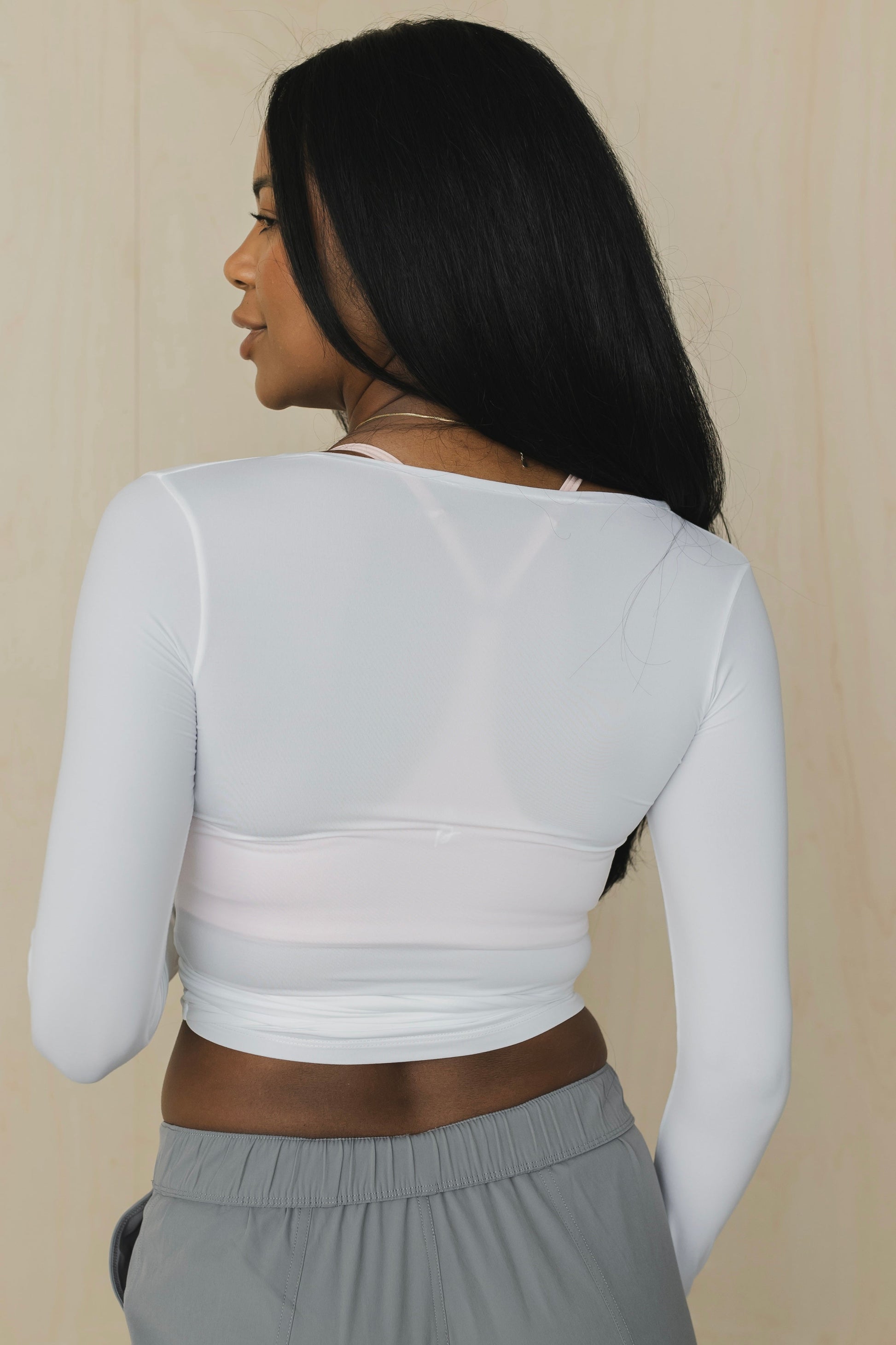 back view of a woman wearing a white long-sleeve top over a pink bralette with gray pants against a beige background
