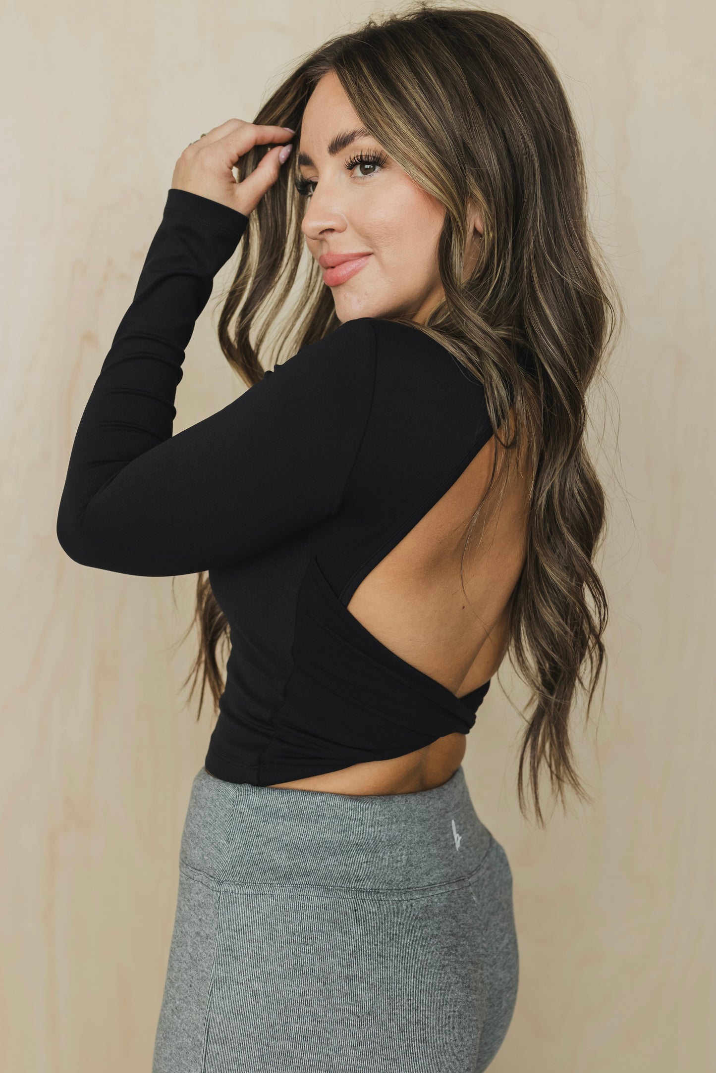 side view of a woman wearing a black long-sleeve crop top with open back and gray leggings against a beige background