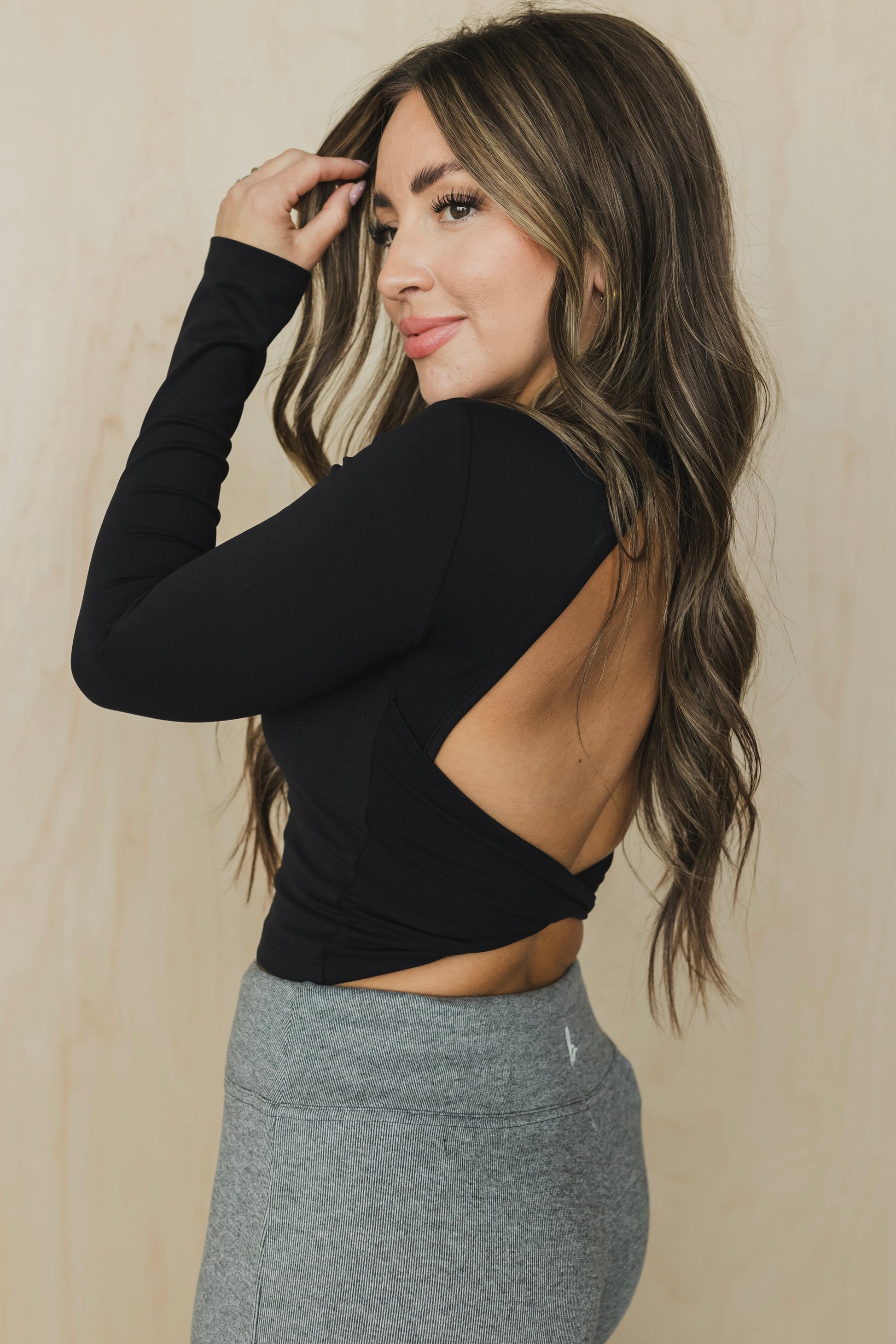 side view of a woman wearing a black long-sleeve crop top with open back and gray leggings against a beige background