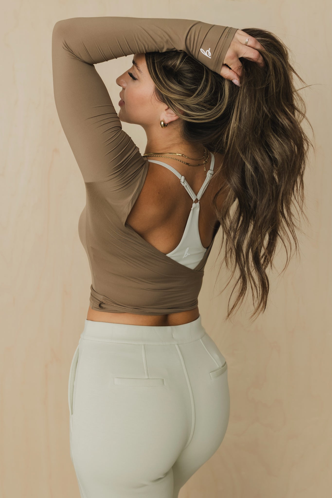 back view of a woman wearing a light brown long-sleeve open back top over a white sports bra with gray pants against a beige background