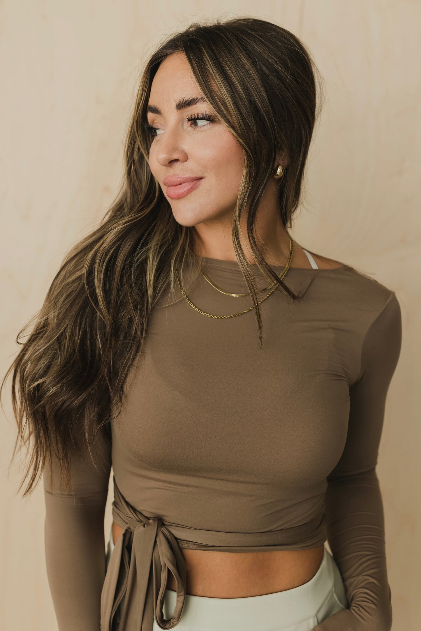 front view of a smiling woman wearing a light brown long-sleeve top with gray pants against a beige background