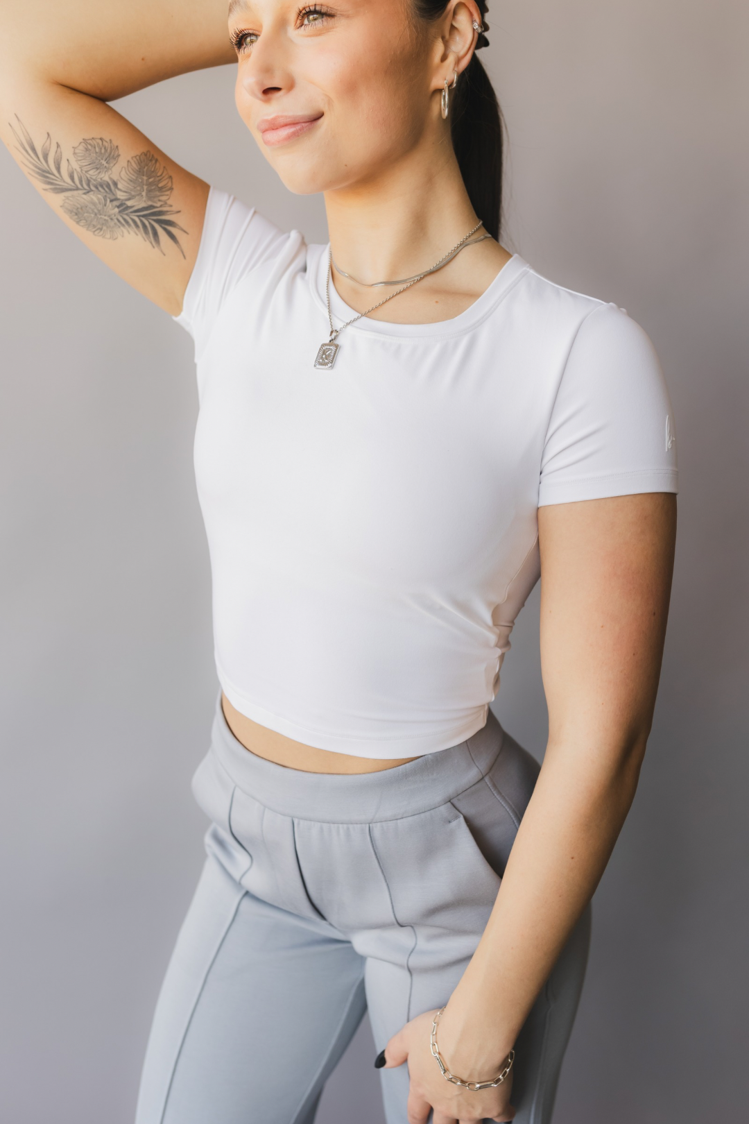 front and side view of a woman wearing a white crop top with cut-out design and light gray pants against a beige background