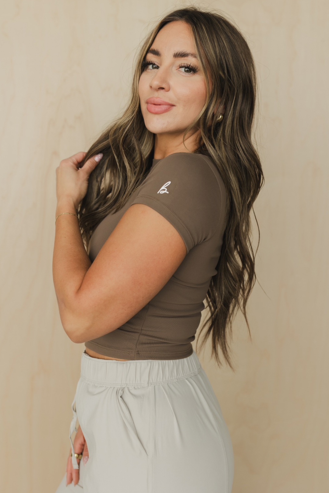 side view of a woman wearing a brown crop top with a logo against a beige background