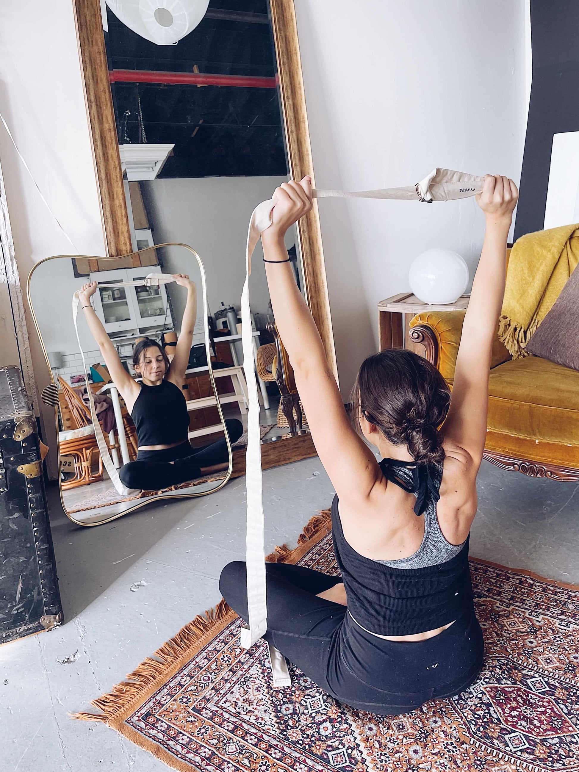 Woman practicing yoga with a yoga strap in a room with a mirror and furniture.