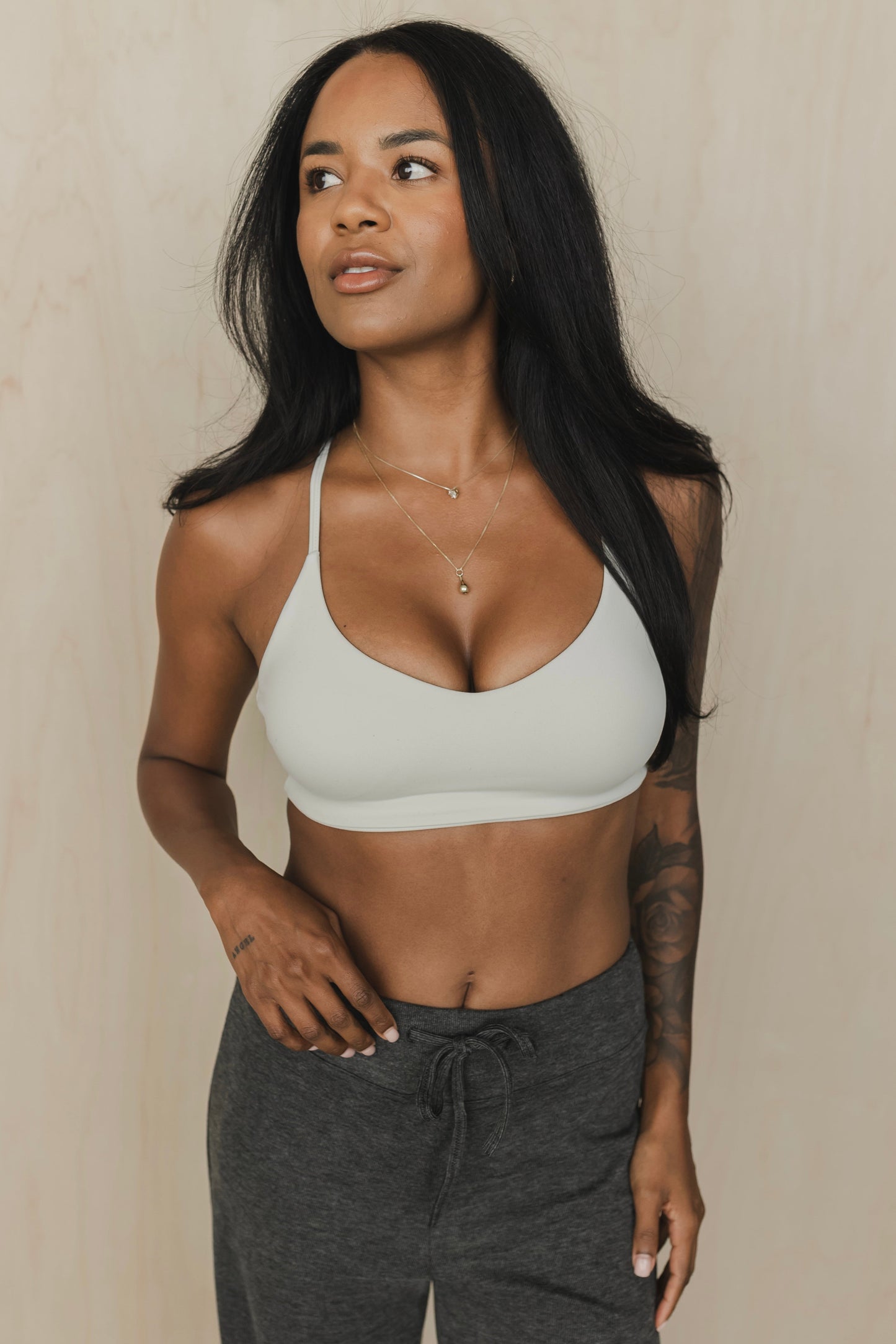 Woman wearing a greyish white sports bra and dark gray sweatpants against a beige background