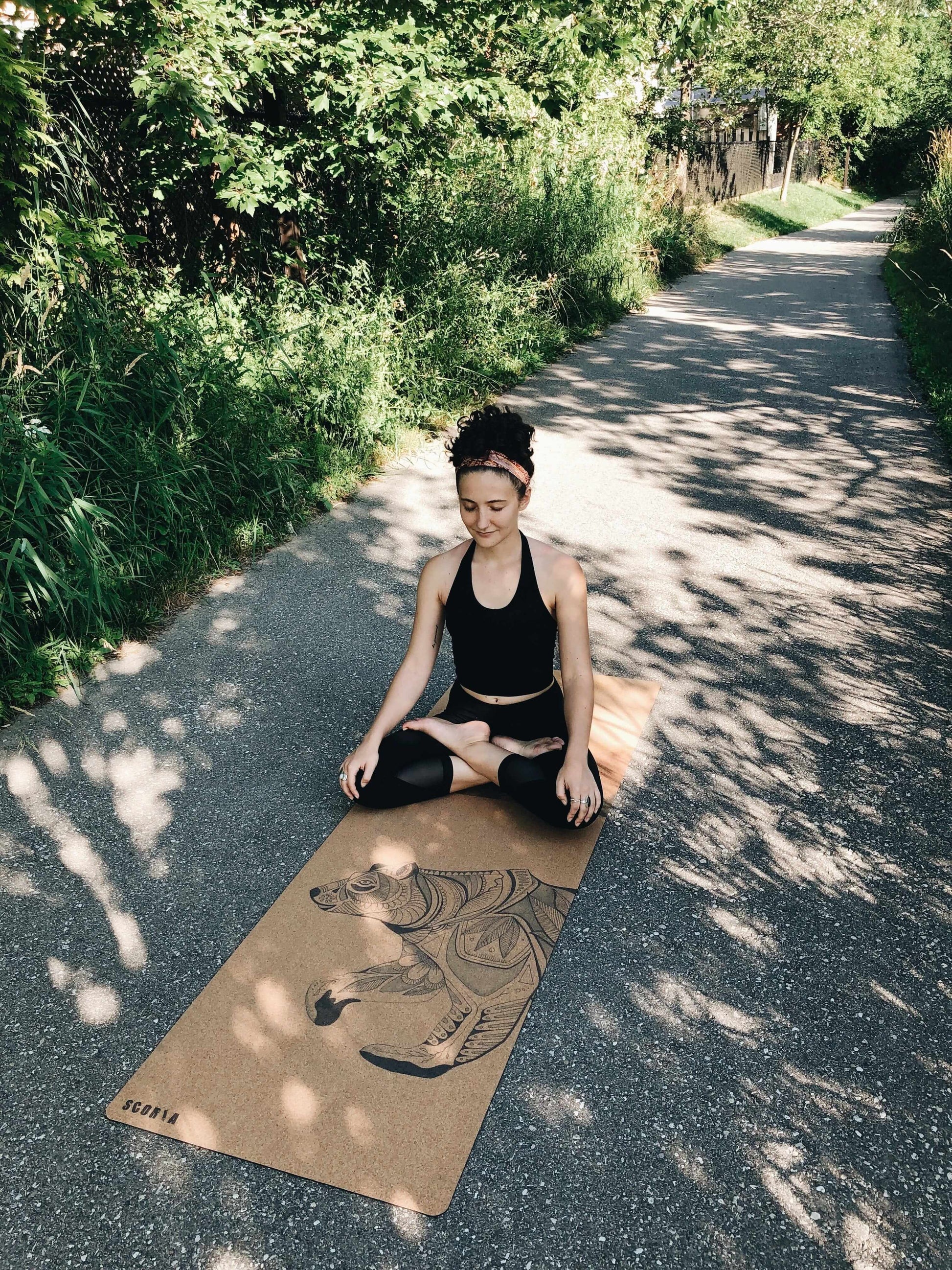 woman sitting on a cork yoga mat with bear design outside on a walking trail