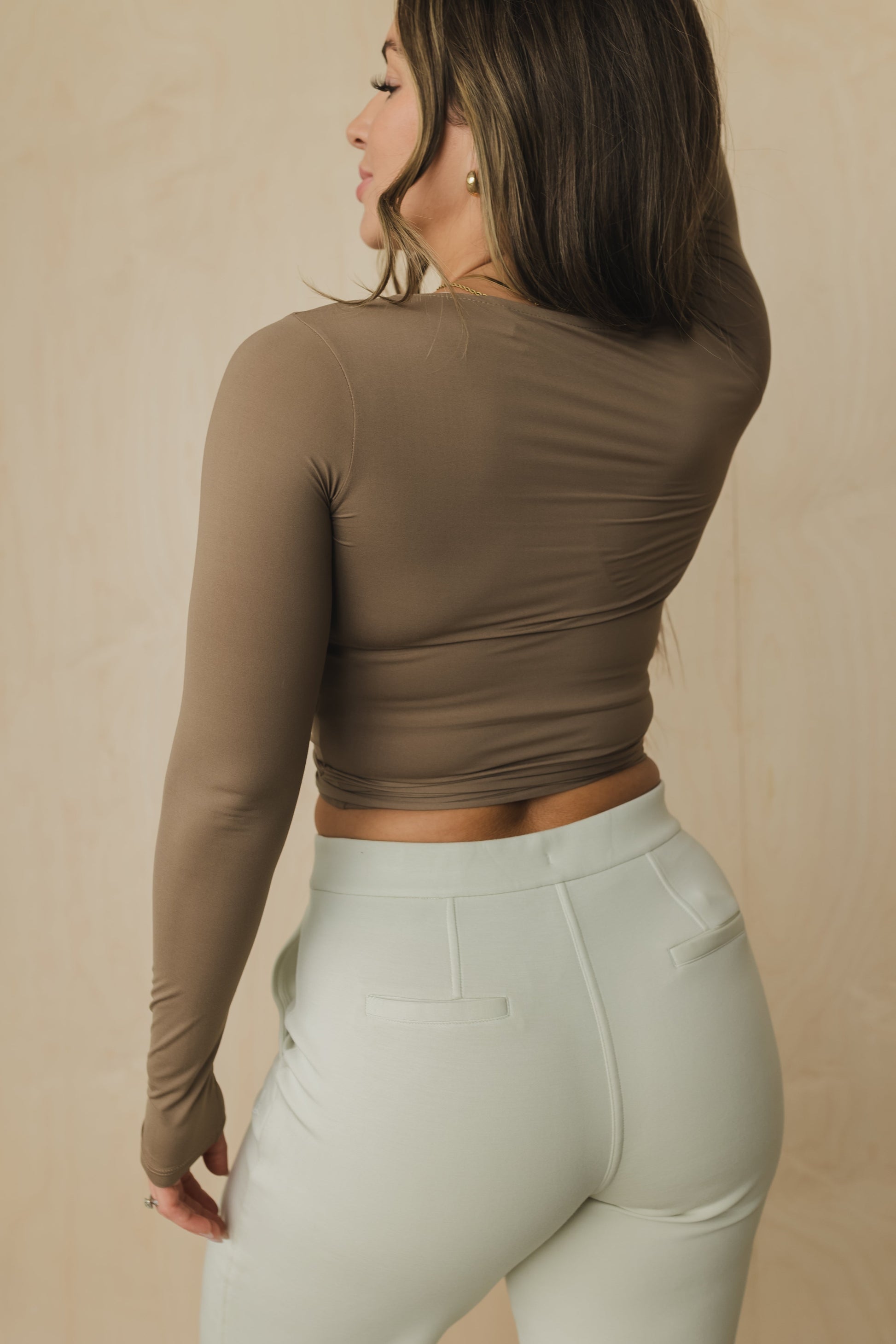 Woman wearing a brown long-sleeve top and light gray pants against a beige background