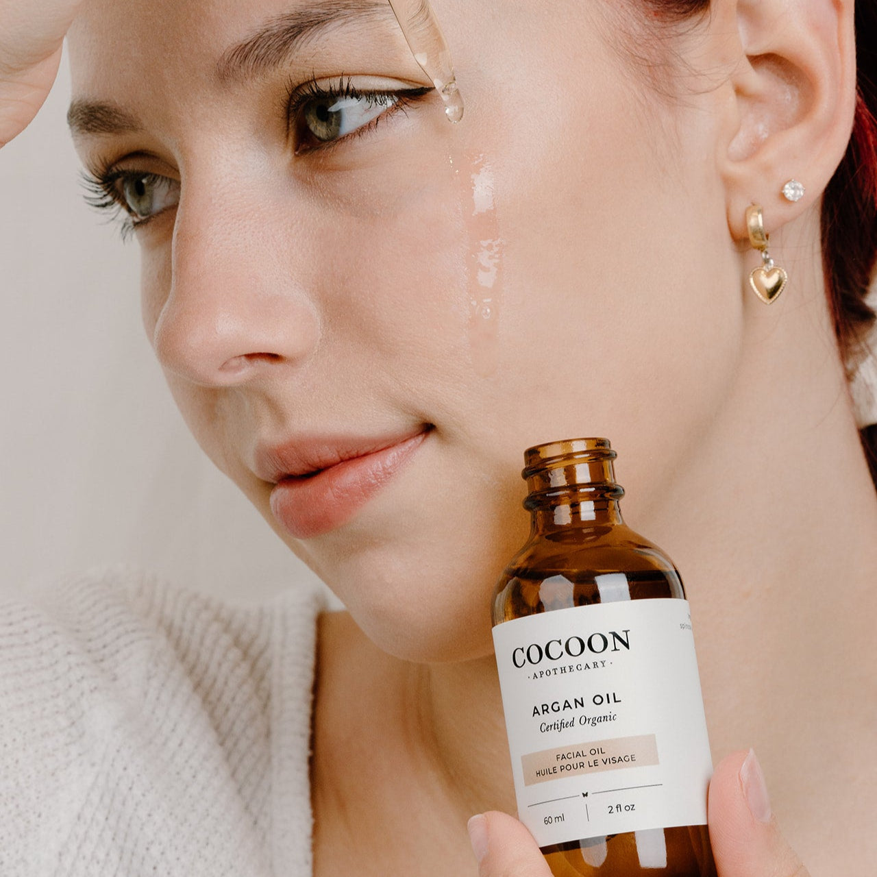 Woman holding a bottle of Cocoon Argan Facial Oil with a close-up of her face.