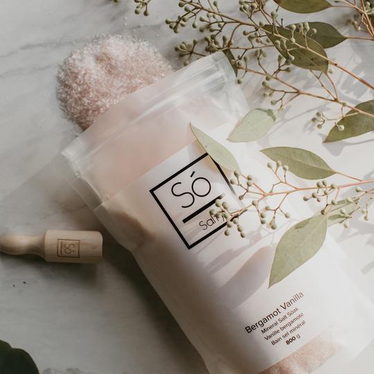 Bag of Bergamot Vanilla Só Salty Bath Salts with a wooden scoop and eucalyptus leaves on a light background