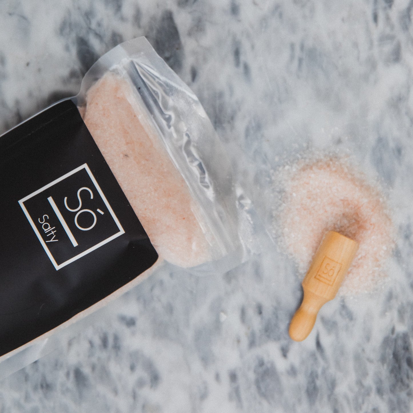 Container of pink bath bomb with a wooden roller on a marble surface, featuring 'So Salty' branding.