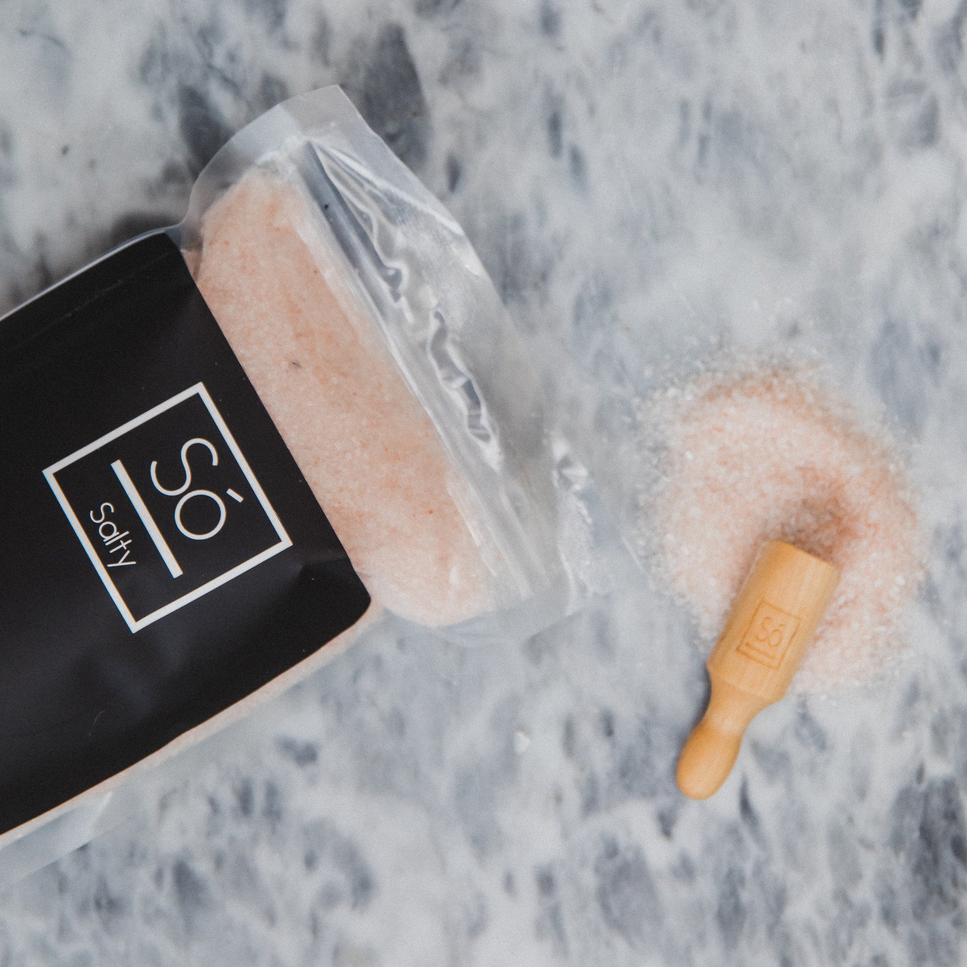 Container of pink bath bomb with a wooden roller on a marble surface, featuring 'So Salty' branding.