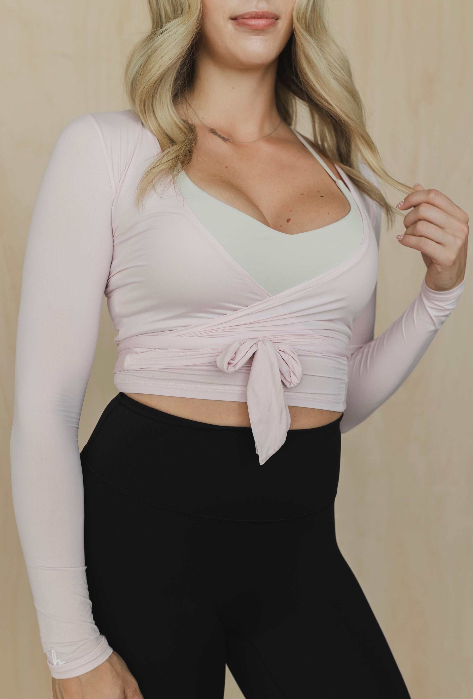Woman wearing a light pink long-sleeve crop top with a tie detail and black leggings against a beige background.