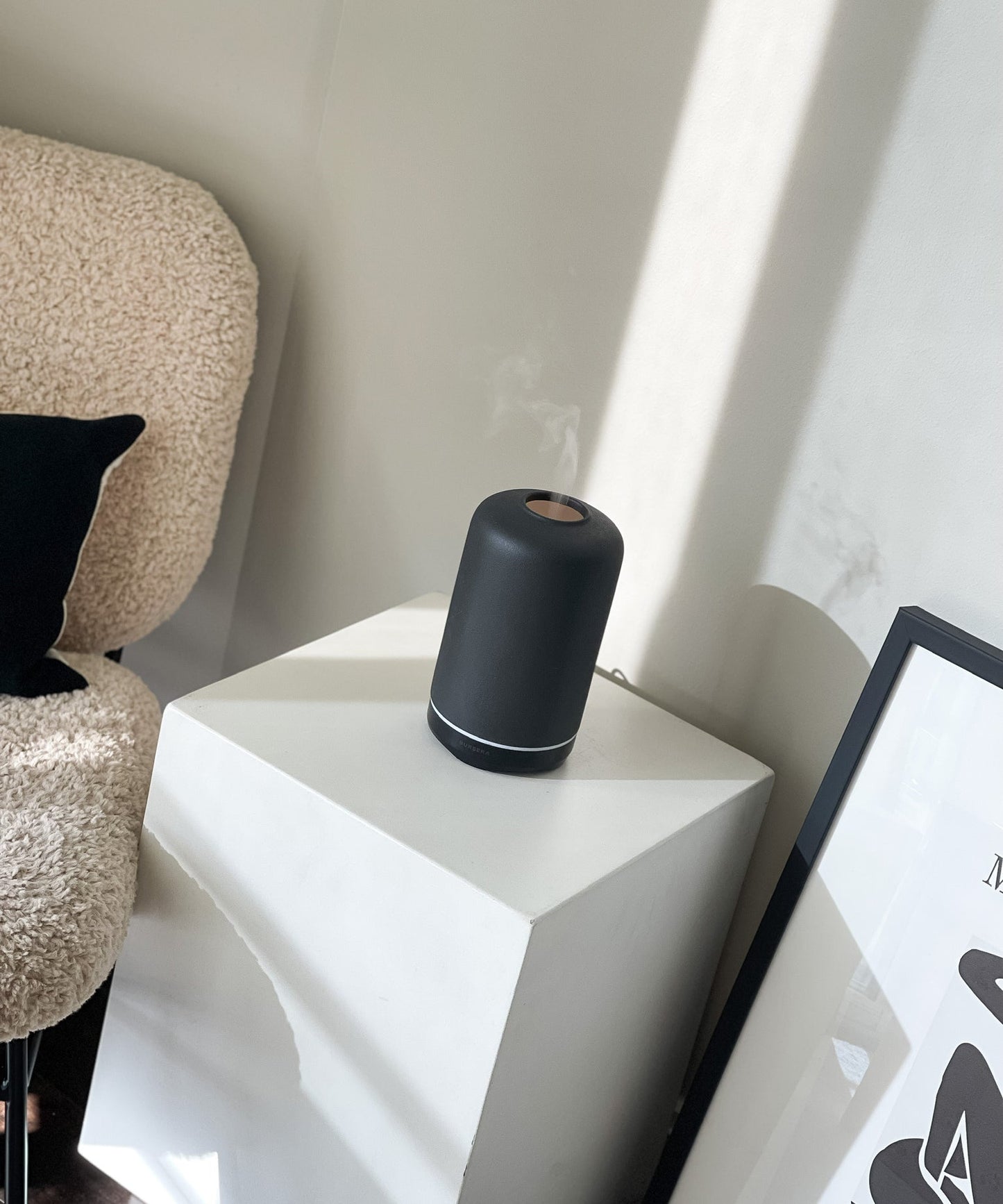 ash essential oil diffuser aromatherapy home decor on a white geometric side table with a textured chair and framed picture in the background.