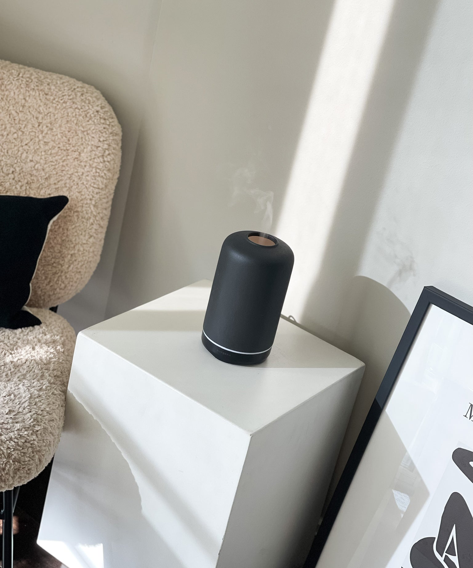 ash essential oil diffuser aromatherapy home decor on a white geometric side table with a textured chair and framed picture in the background.