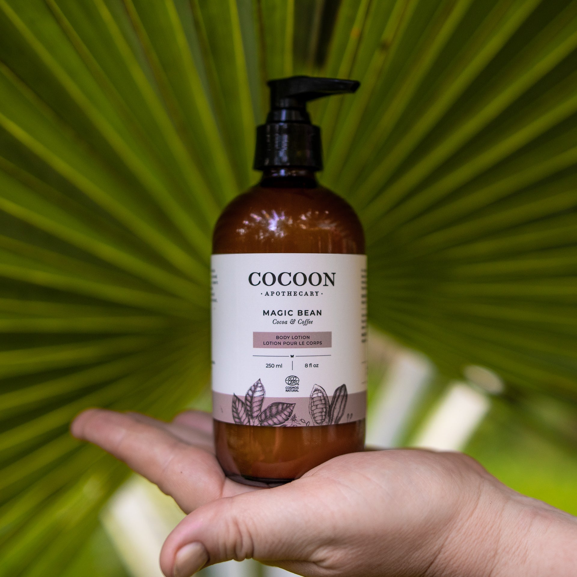 Cocoon Apothecary Magic Bean lotion bottle held in a hand with a green leafy background