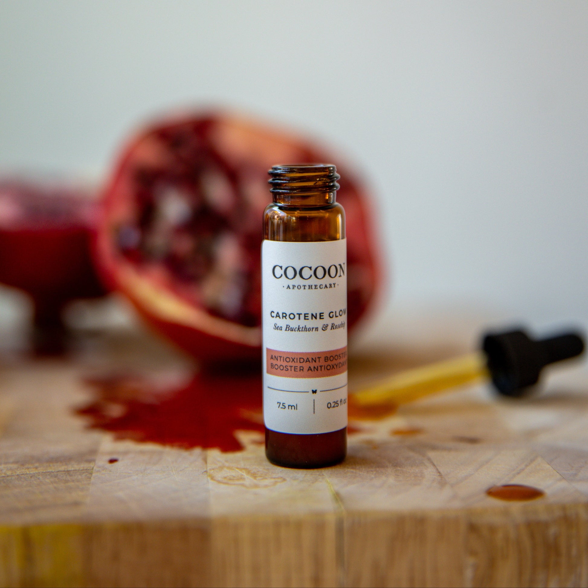 Small bottle of Carotene Glow Antioxidant Booster labeled 'Cocoon' on a wooden surface with a pomegranate in the background.