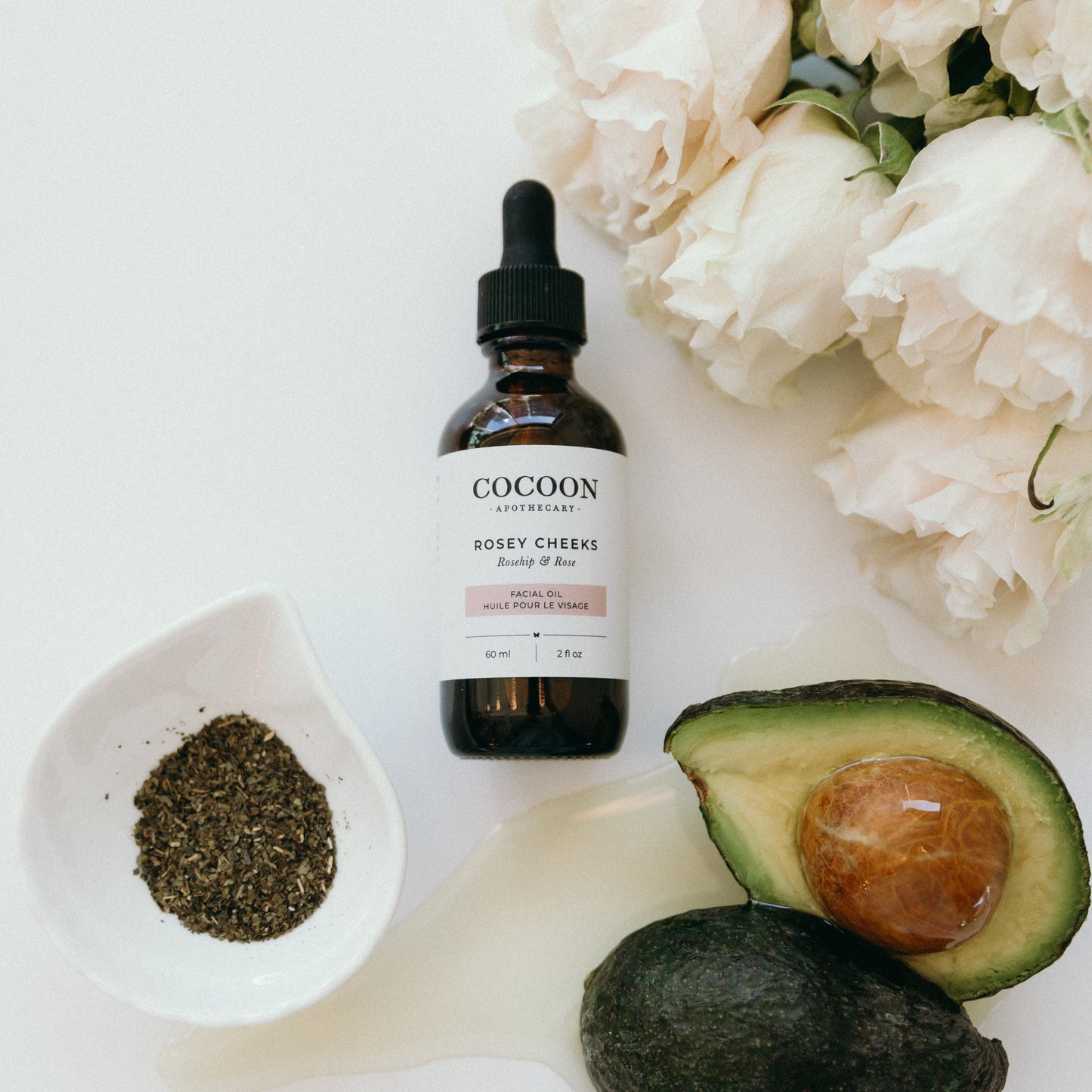 Cocoon skincare facial oil bottle with avocado, flowers, and a spoon on a white background