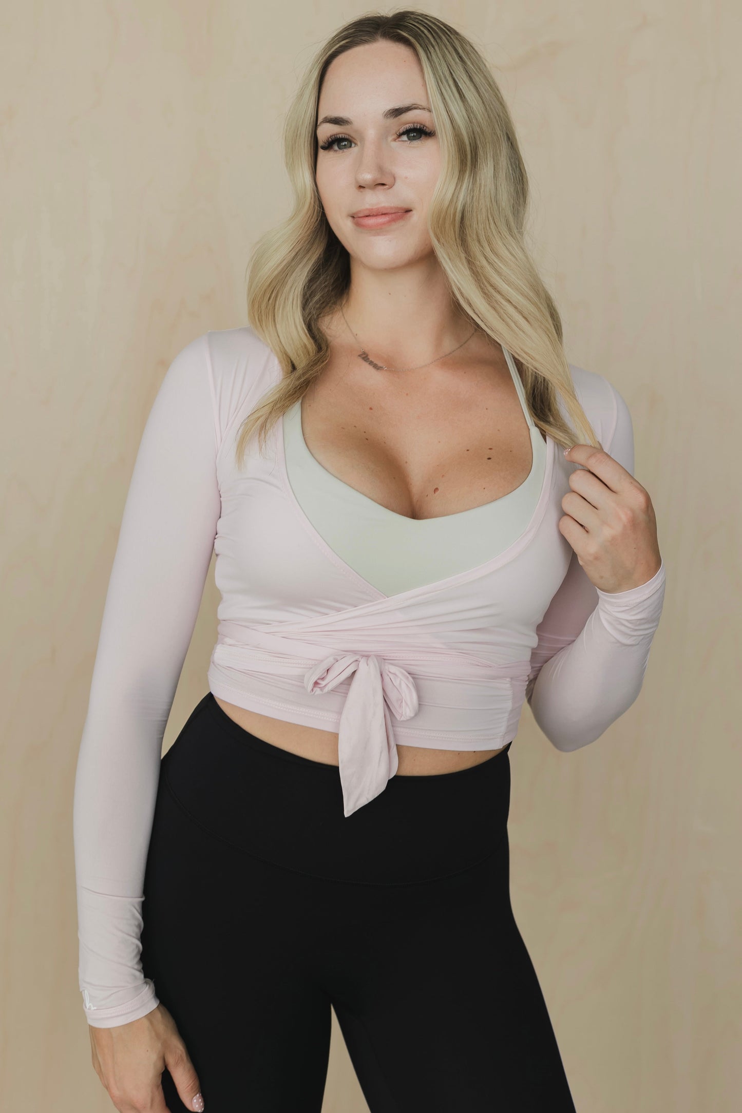 Woman wearing a light pink long-sleeve top with a tie detail over a green top and black pants on a beige background.