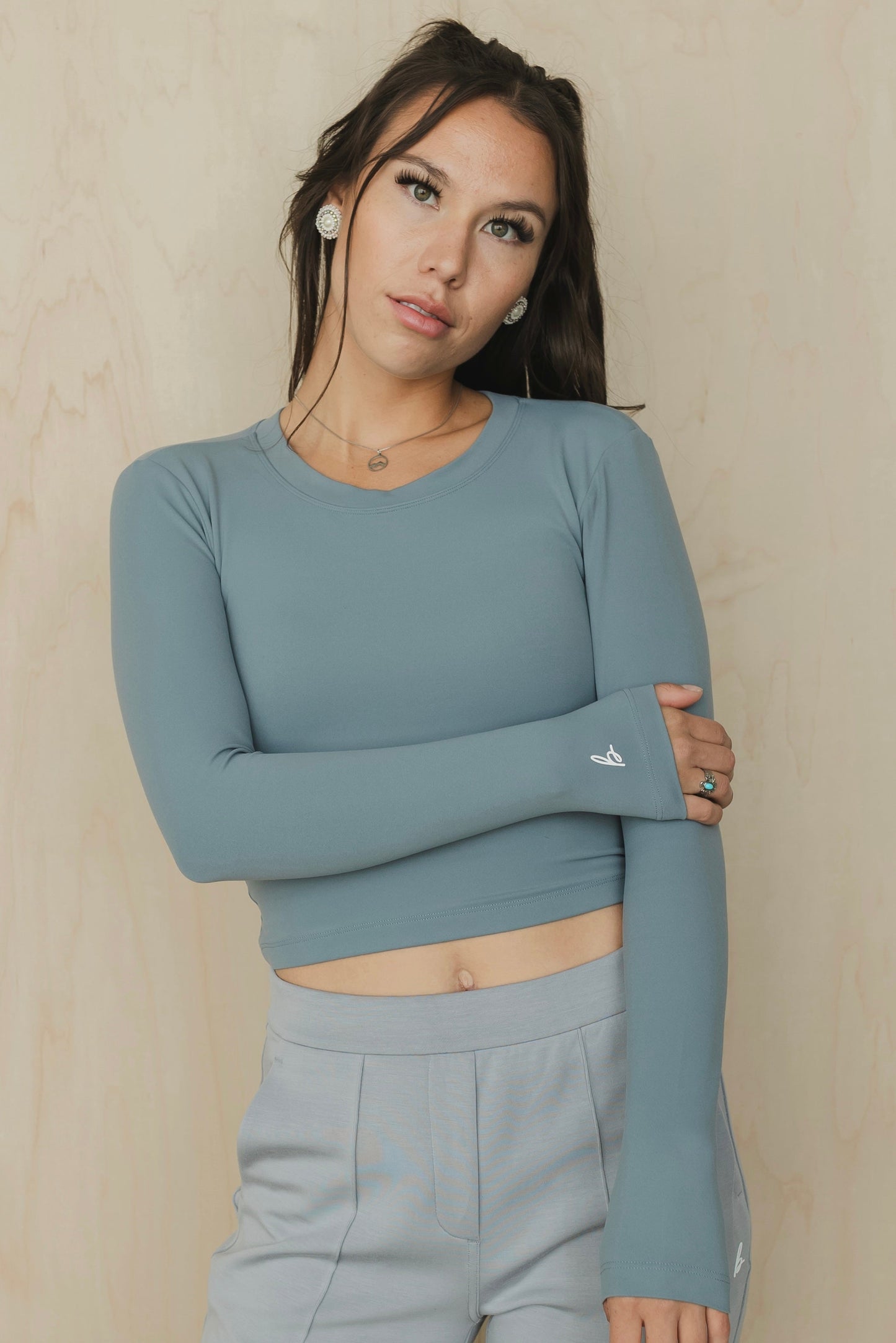 front cose up view of a woman wearing a teal long-sleeve backless top with a brand logo against a beige background