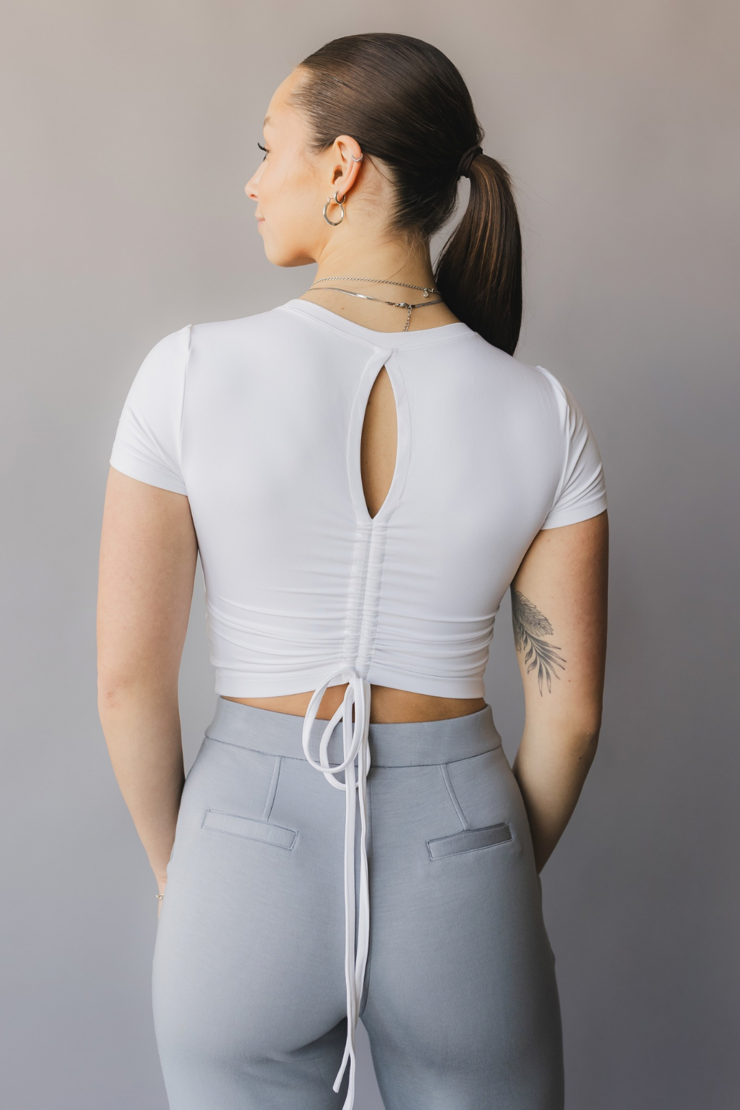 back view of a woman wearing a white crop top with cut-out design and light gray pants against a beige background