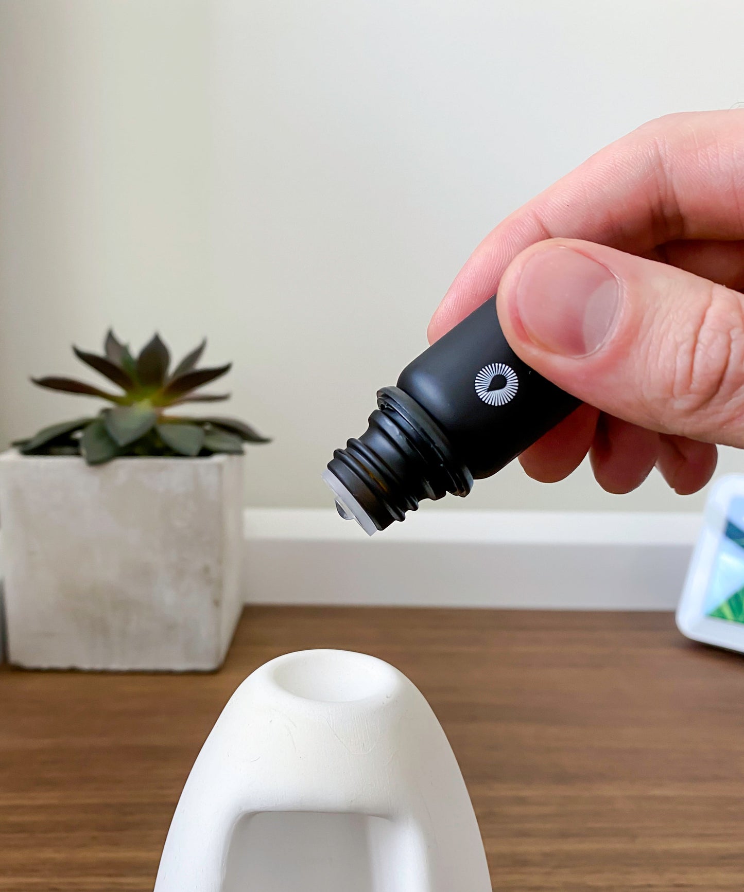 Hand holding a black essential oil dropper near a focus diffuser kit essential oil aromatherapy set on a wooden surface with a plant in the background.