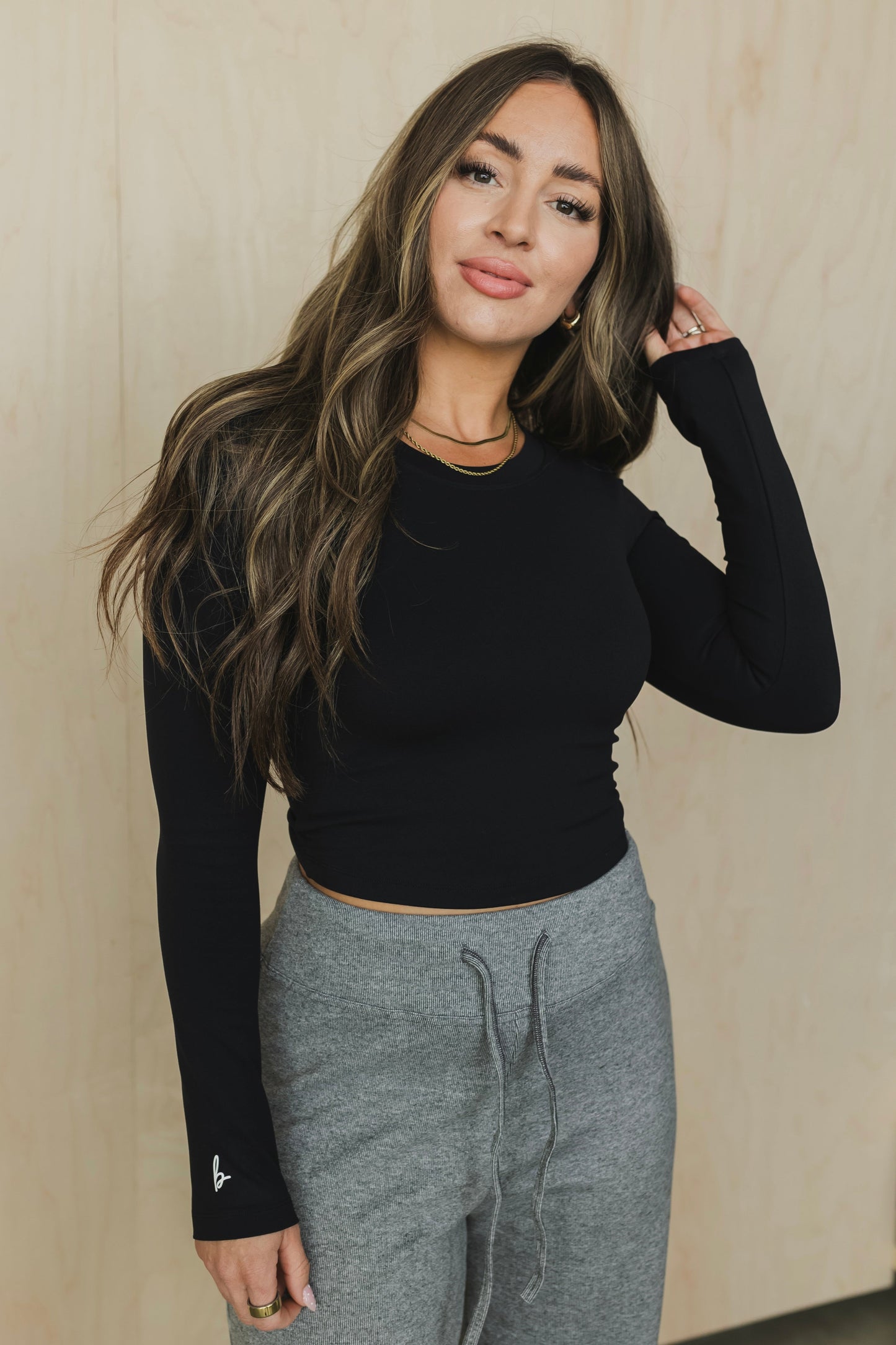 front view of a woman wearing a black long-sleeve open back top and gray sweatpants against a beige wall.