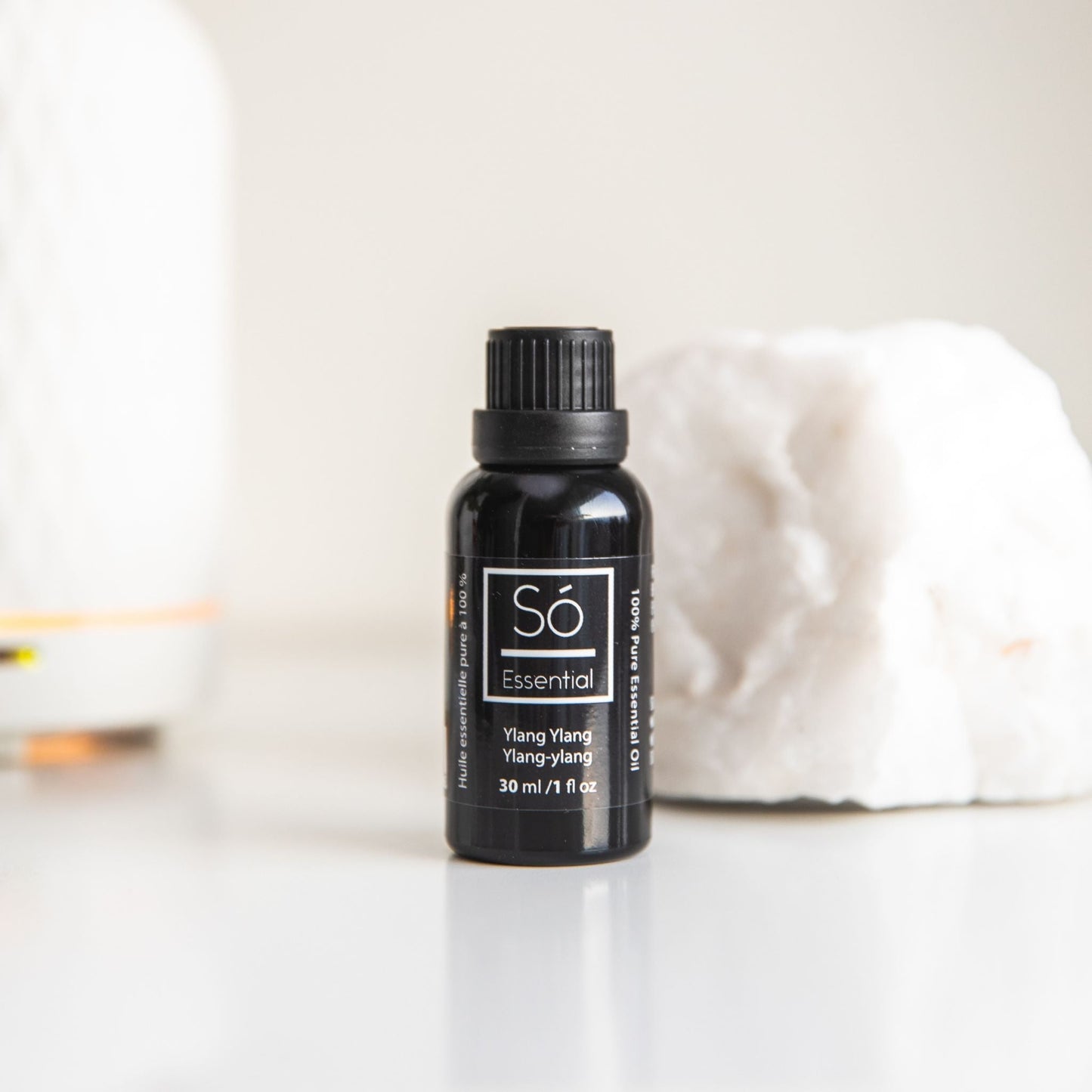 Black Ylang Ylang essential oil bottle labeled 'Sö Essential' on a white surface with a blurred background.