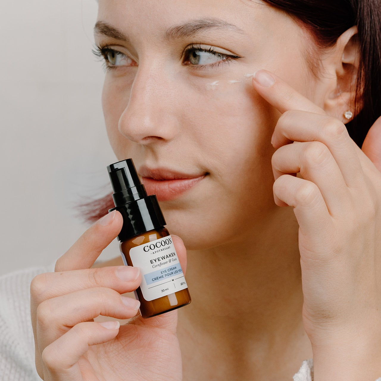 Woman applying 'Eyewaken' eye cream with a bottle labeled 'COCOON' on a neutral background