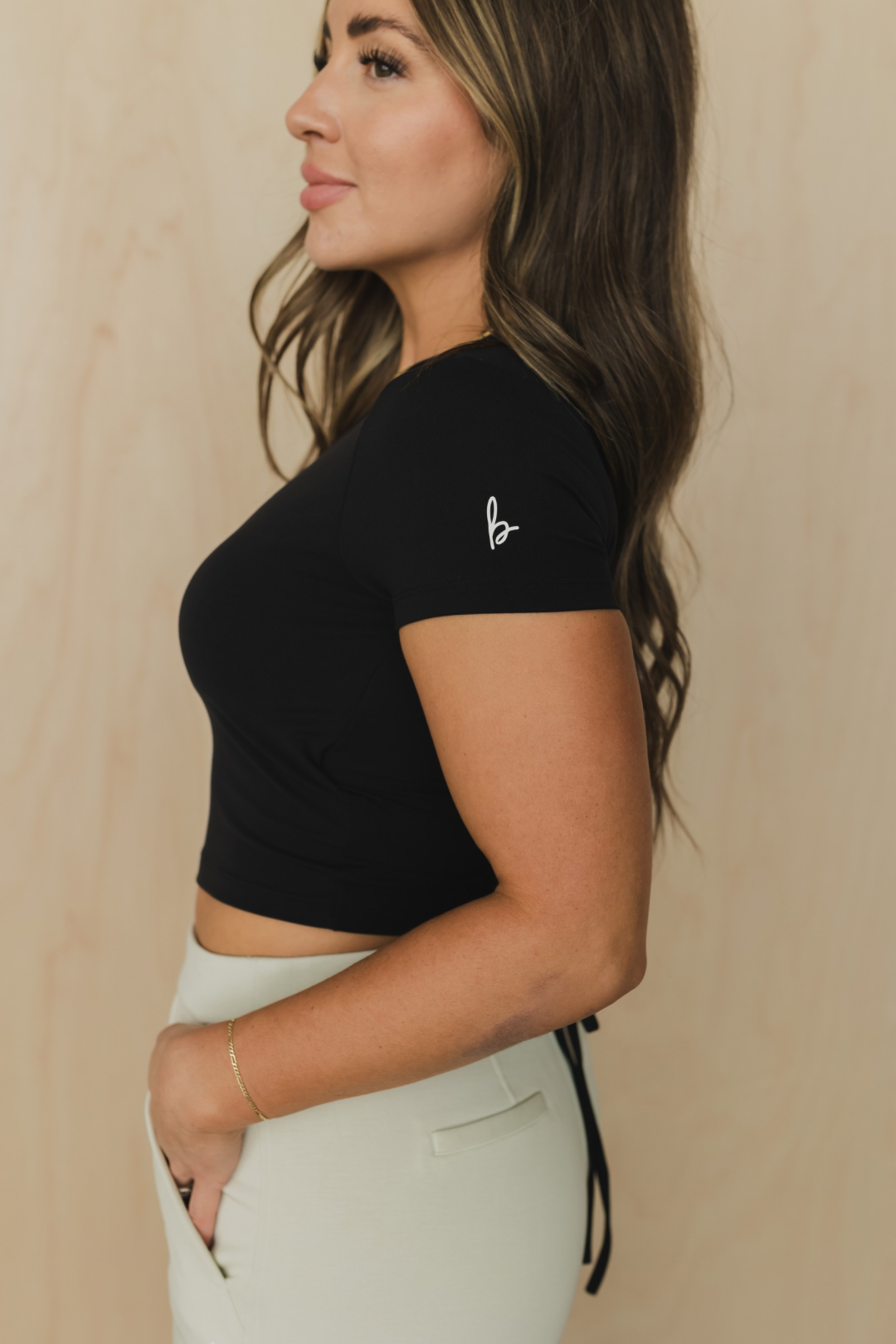 side view of a woman wearing a black crop top with cut-out design with logo and light gray pants against a beige background