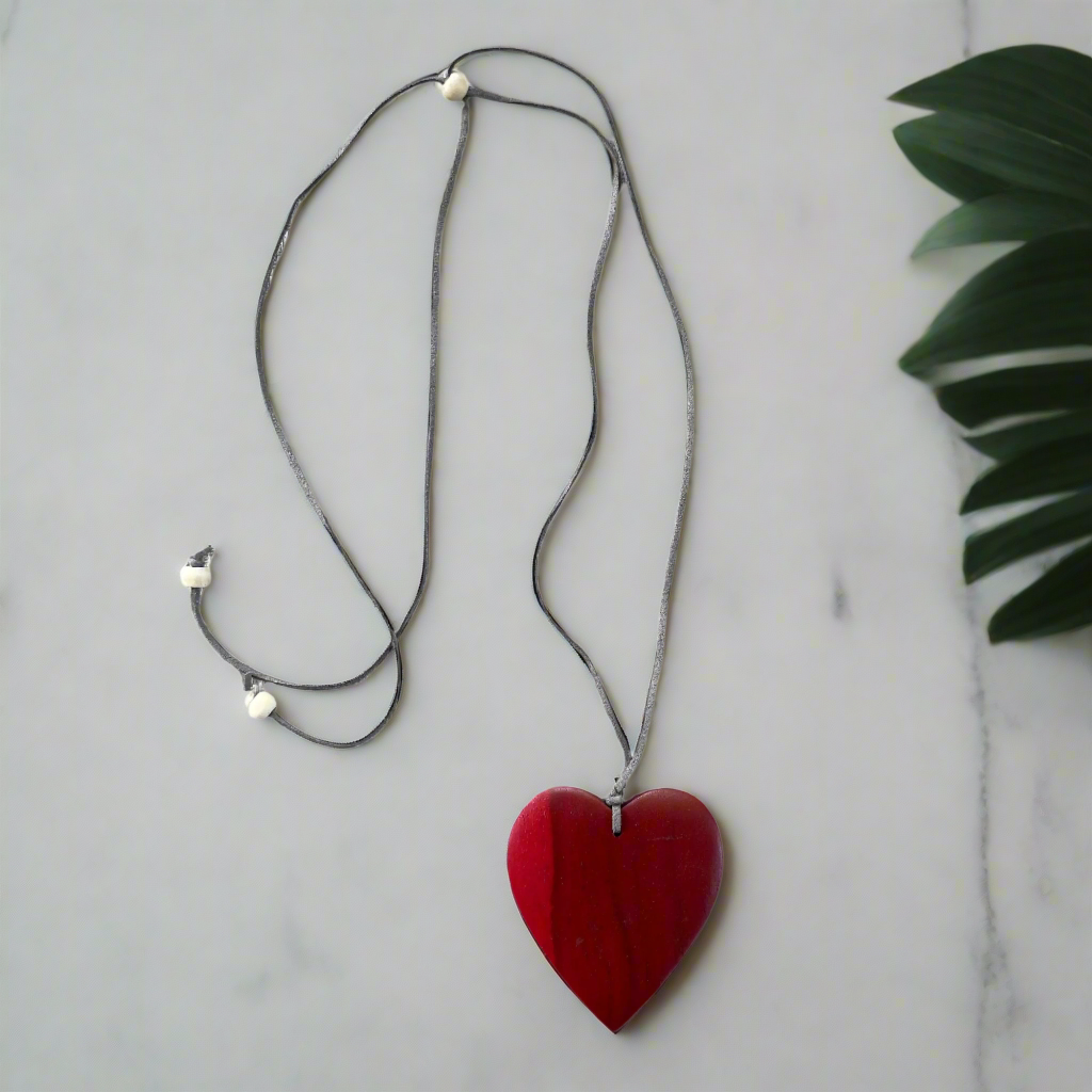 Red heart-shaped pendant on a black cord against a light gray background with a green leaf.