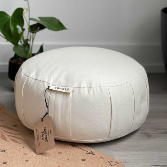 Round white meditation cushion with a tag on a wooden floor next to a plant