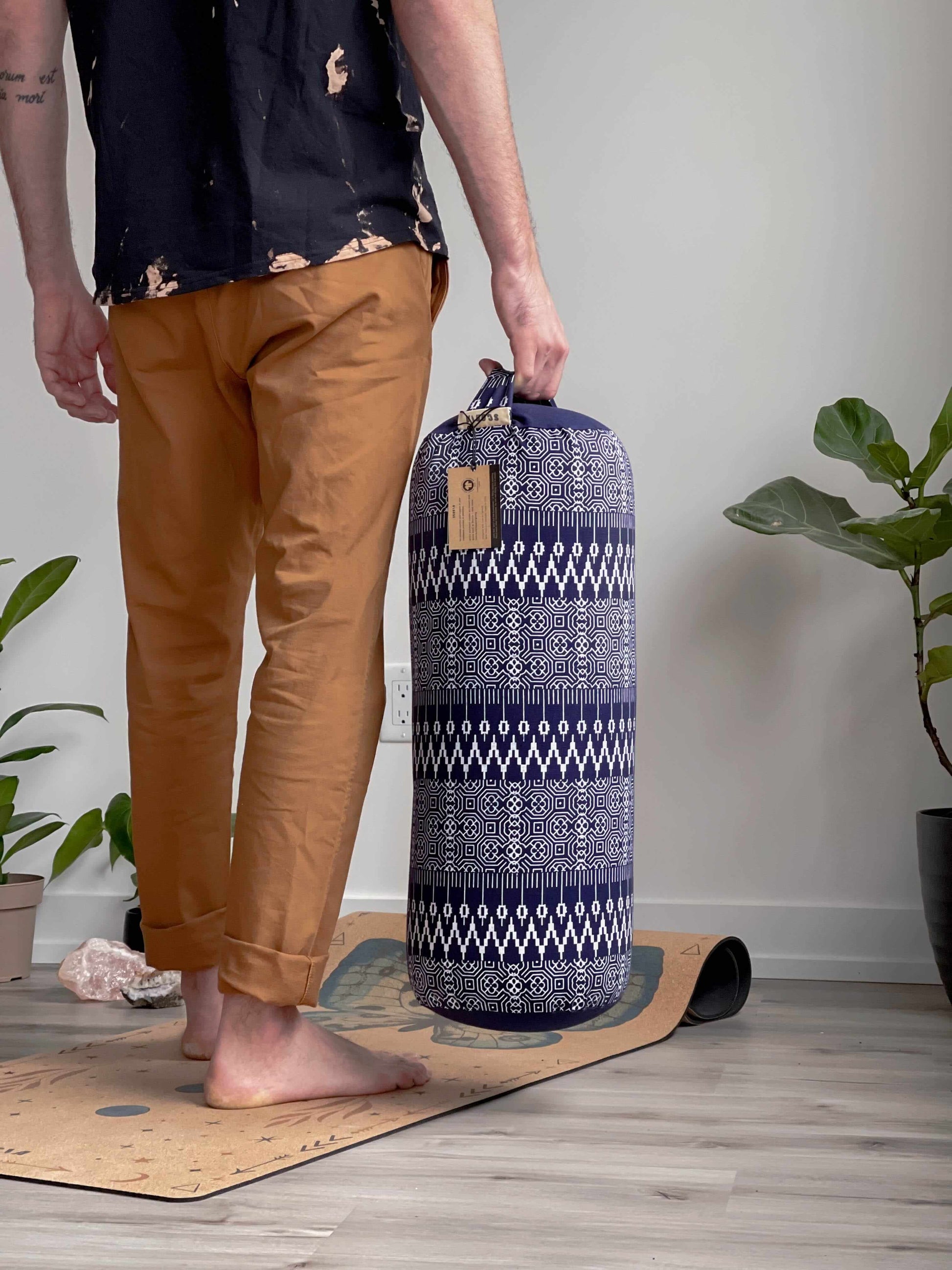 Person holding a patterned yoga mat bag on a wooden floor with plants in the background