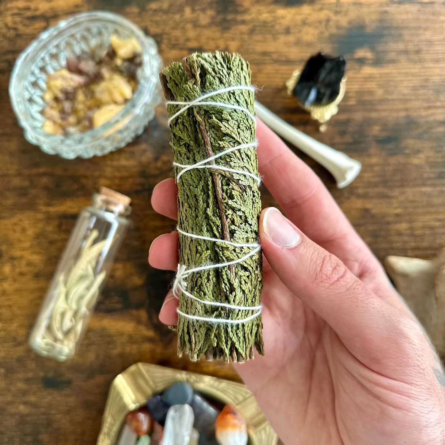 Hand holding a bundle of cedar sage wrapped in twine on a wooden surface with various items.
