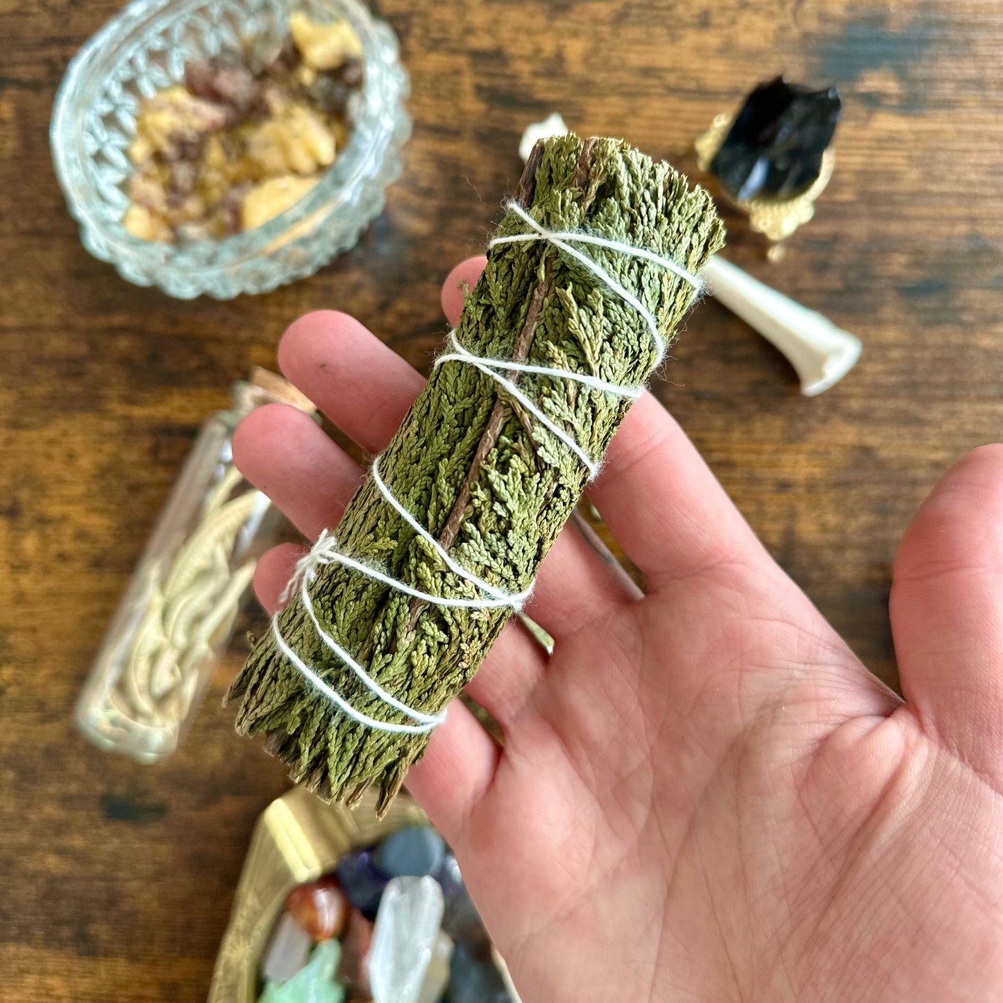 Hand holding a cedar sage bundle wrapped in twine on a wooden surface with various items.
