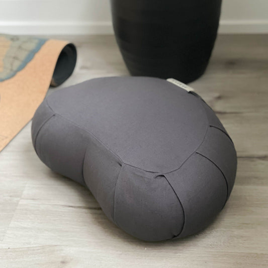 Gray meditation pillow on a wooden floor with a yoga mat and black container in the background.