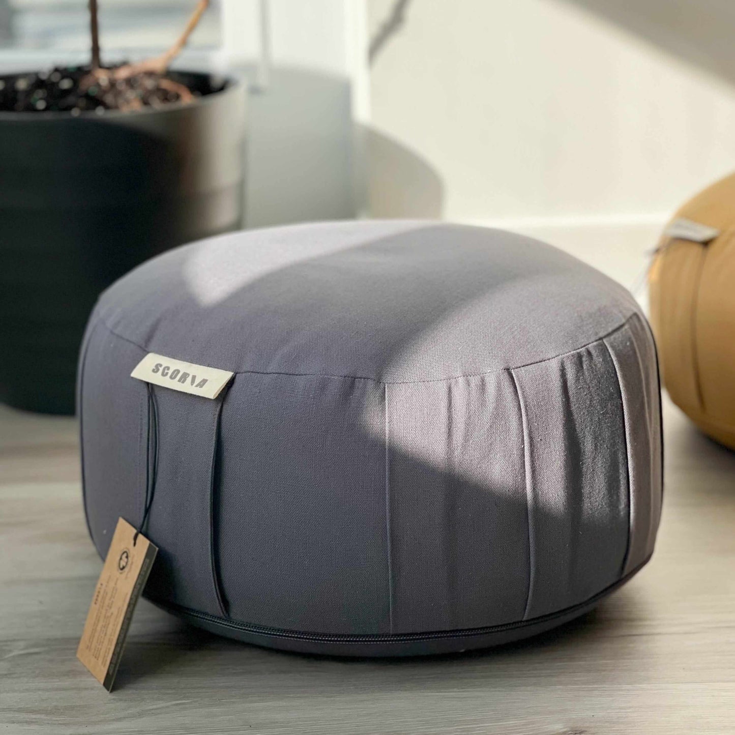 Gray meditation cushion with a tag on a wooden floor, surrounded by indoor plants.