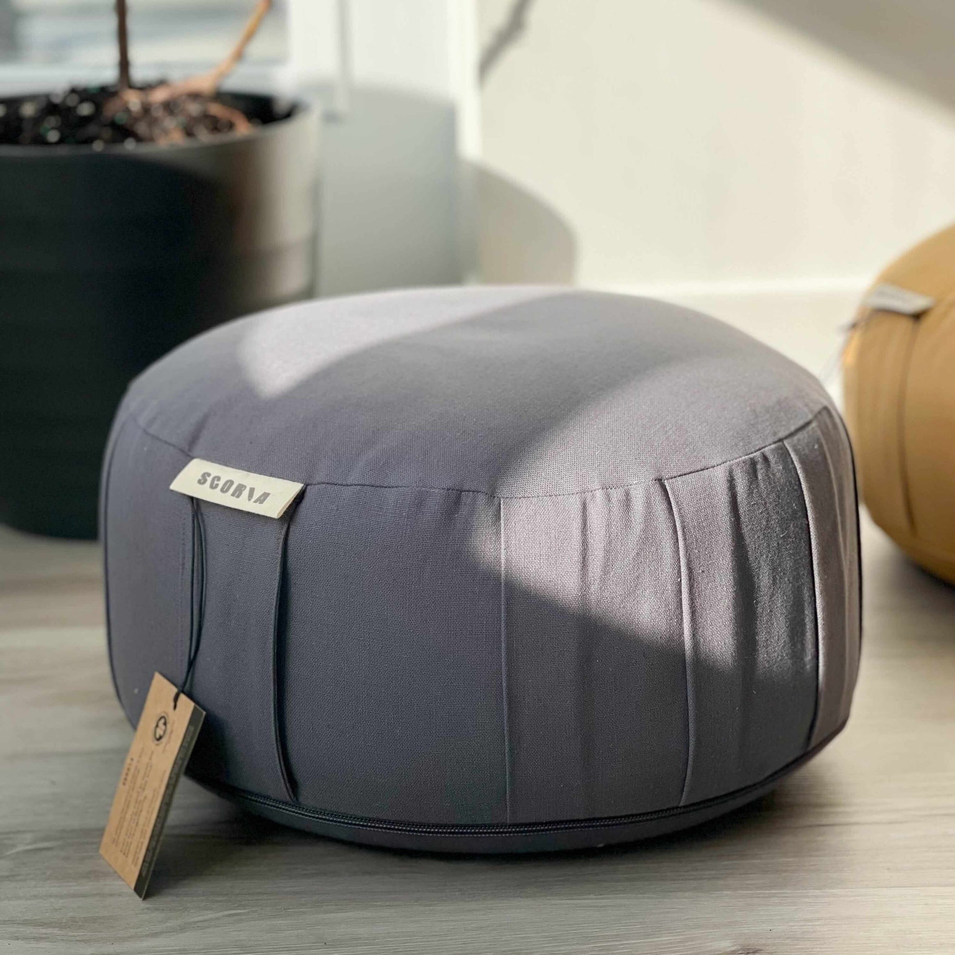 Gray meditation cushion with a tag on a wooden floor, surrounded by indoor plants.