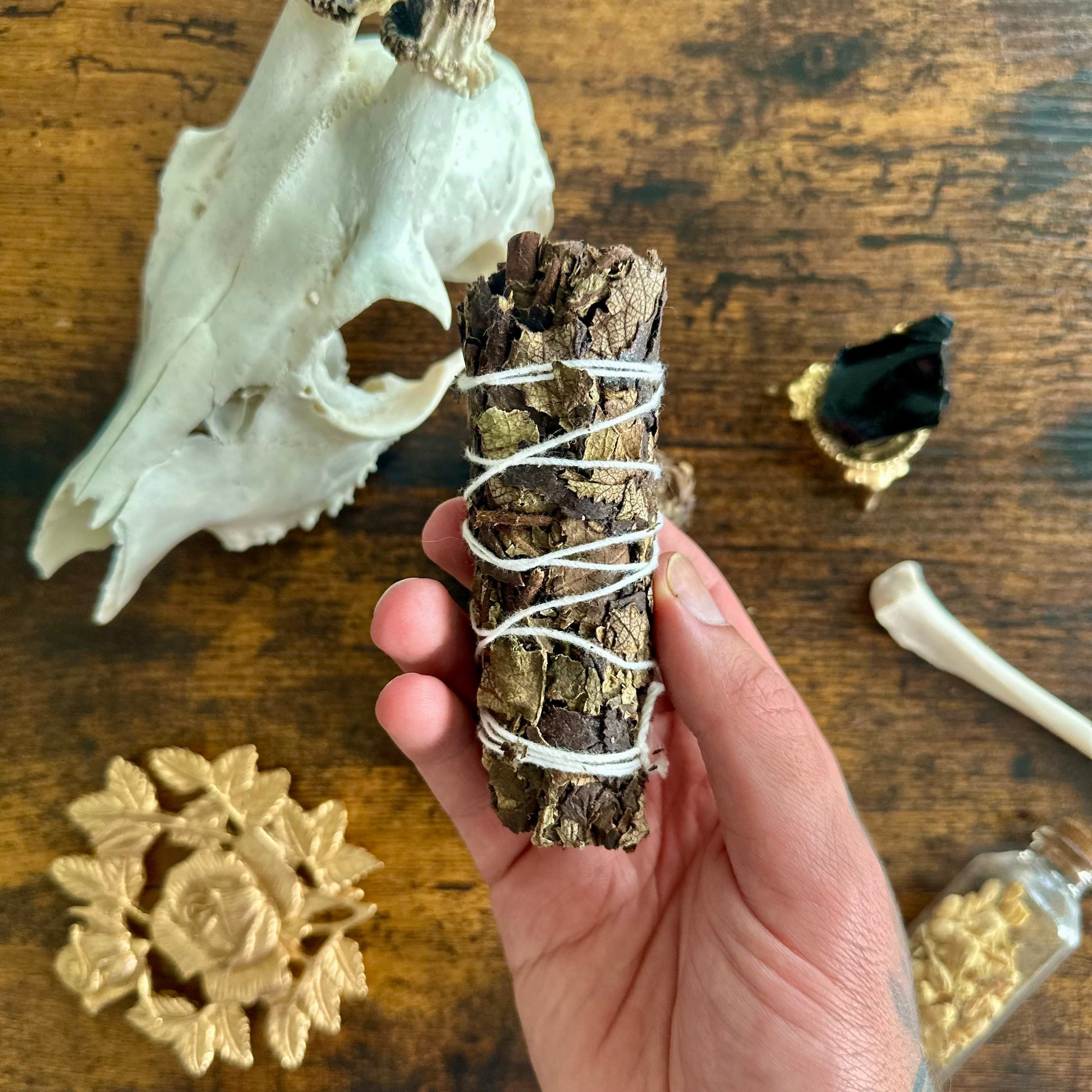Hand holding a bundle of sage with a skull and other items on a wooden surface