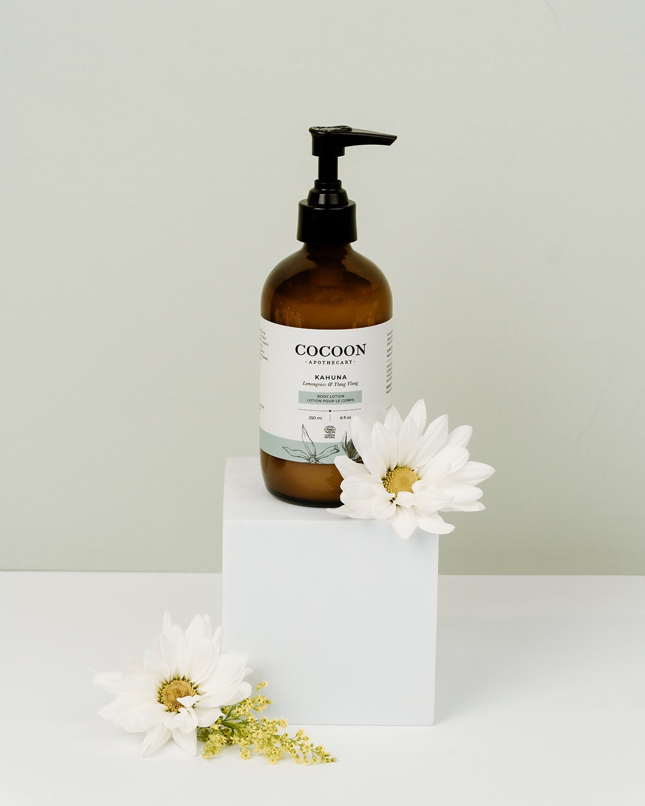Brown bottle of body lotion labeled 'COCOON' with a white label on a white surface with white flowers.
