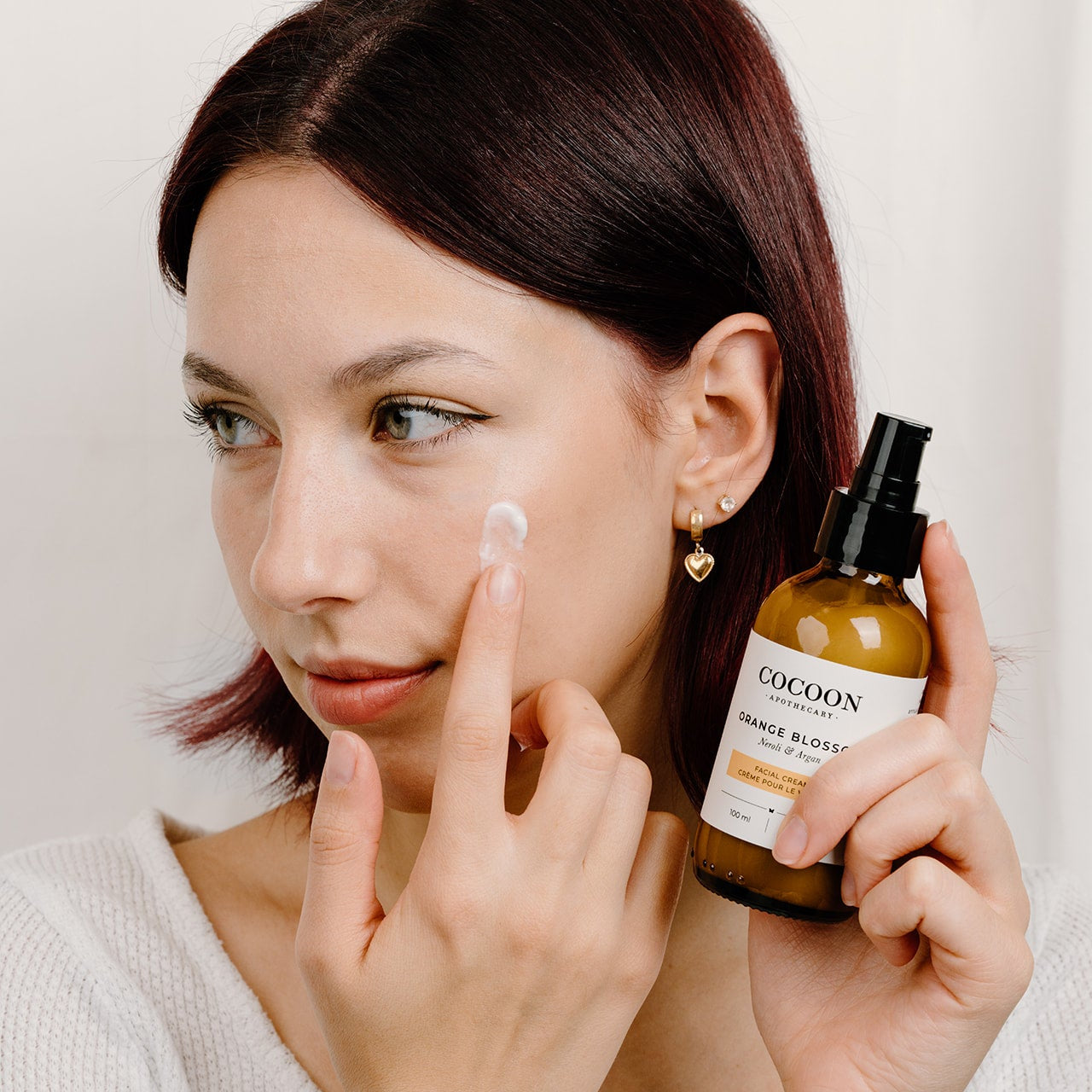 Woman applying a facial cream to her face with a bottle labeled 'Cocoon' in the other hand.