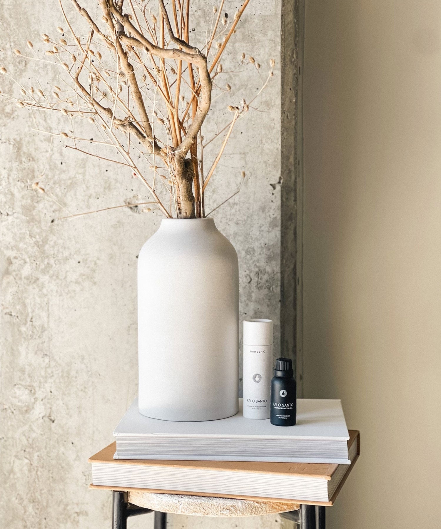 organic palo santo essential oil 30ml aromatherapy beside white vase with dried branches on a wooden surface against a textured wall.