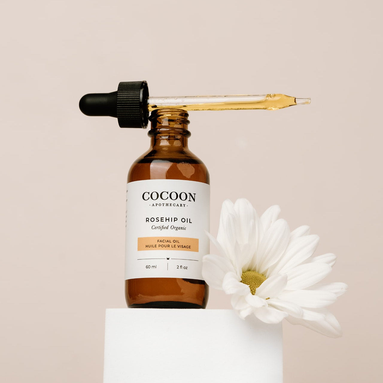 Cocoon Apothecary rosehip oil bottle with dropper and white flower on beige background