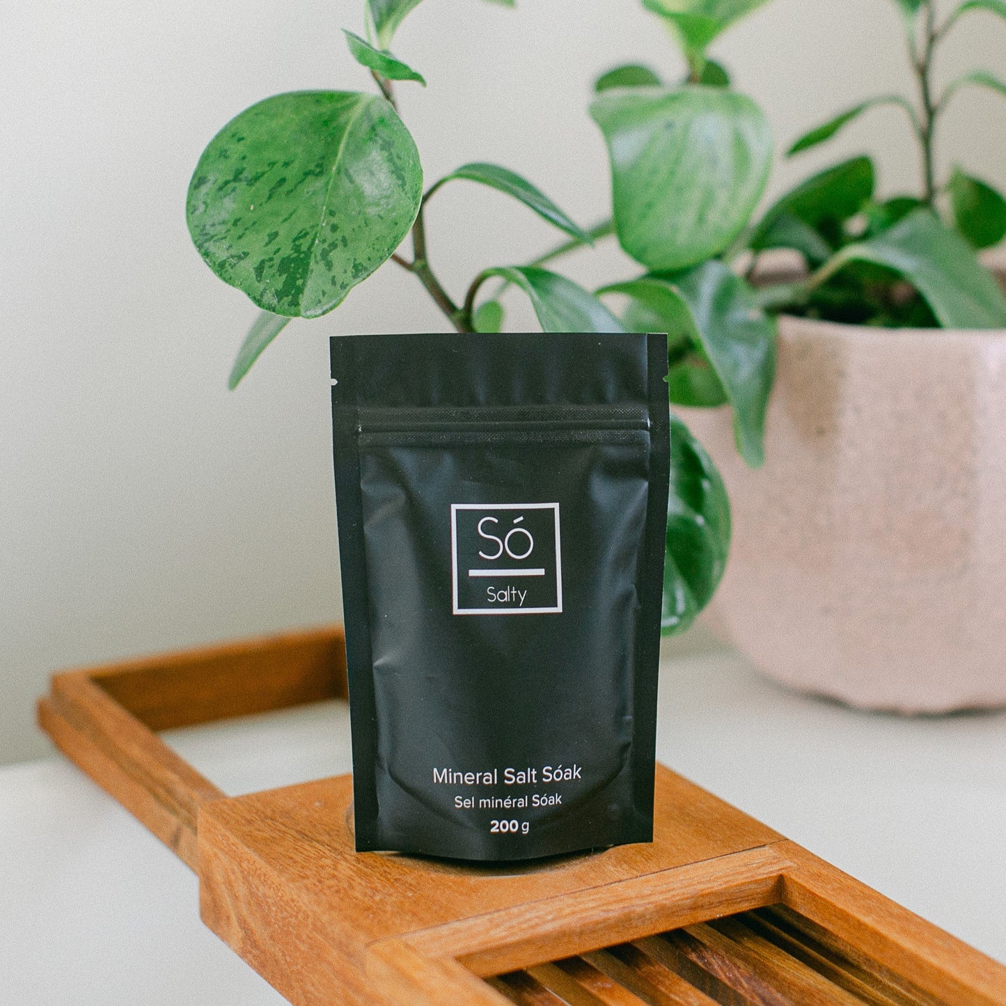Black package of bath salts labeled 'Sö Salty' on a wooden tray with a plant in the background