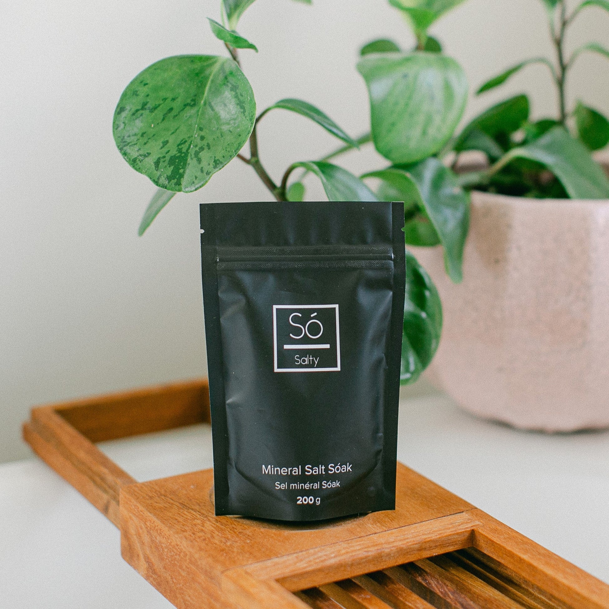Black package of bath salts labeled 'Sö Salty' on a wooden tray with a plant in the background
