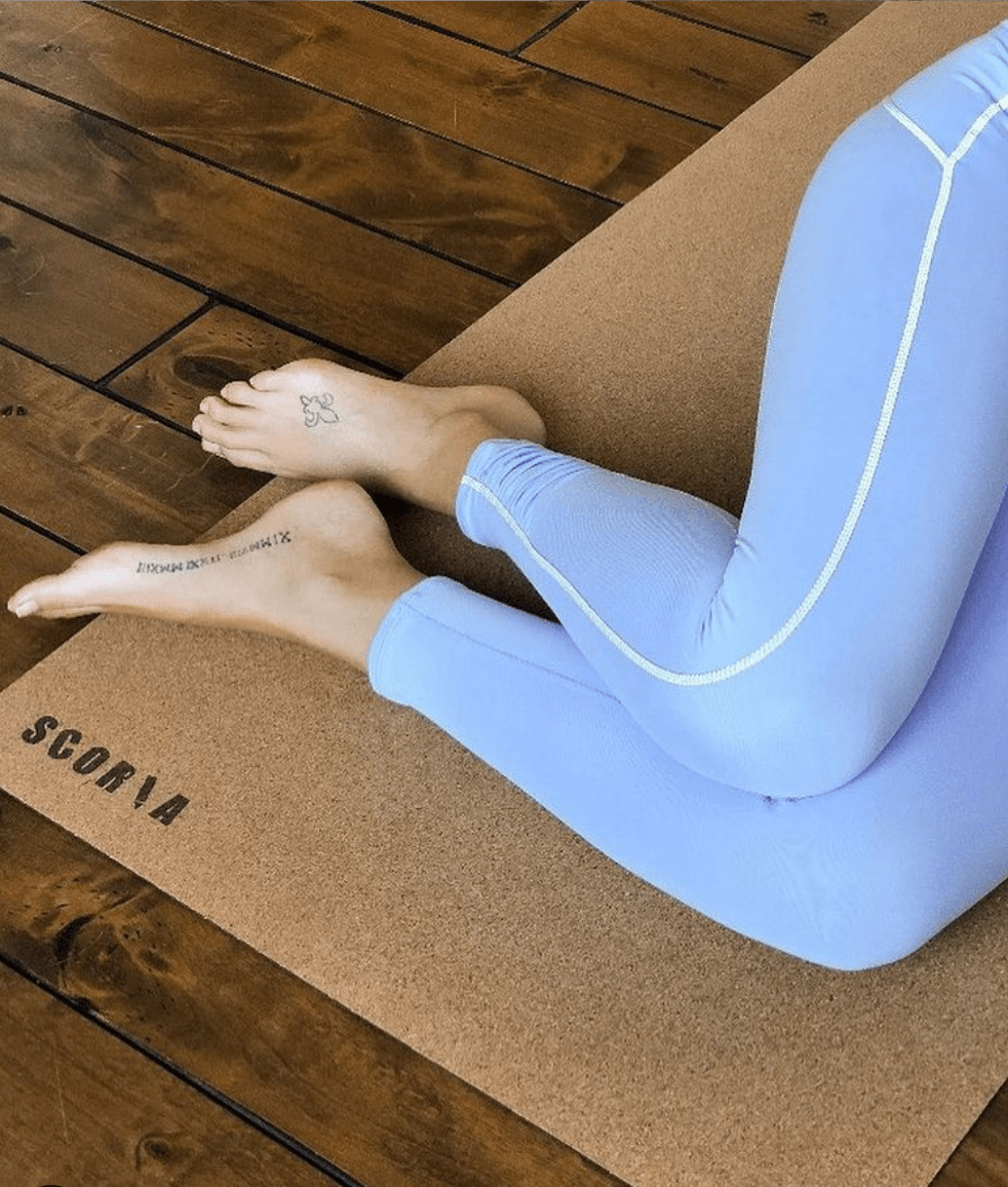 Person wearing light blue leggings sitting on a travel size cork yoga mat with branding.