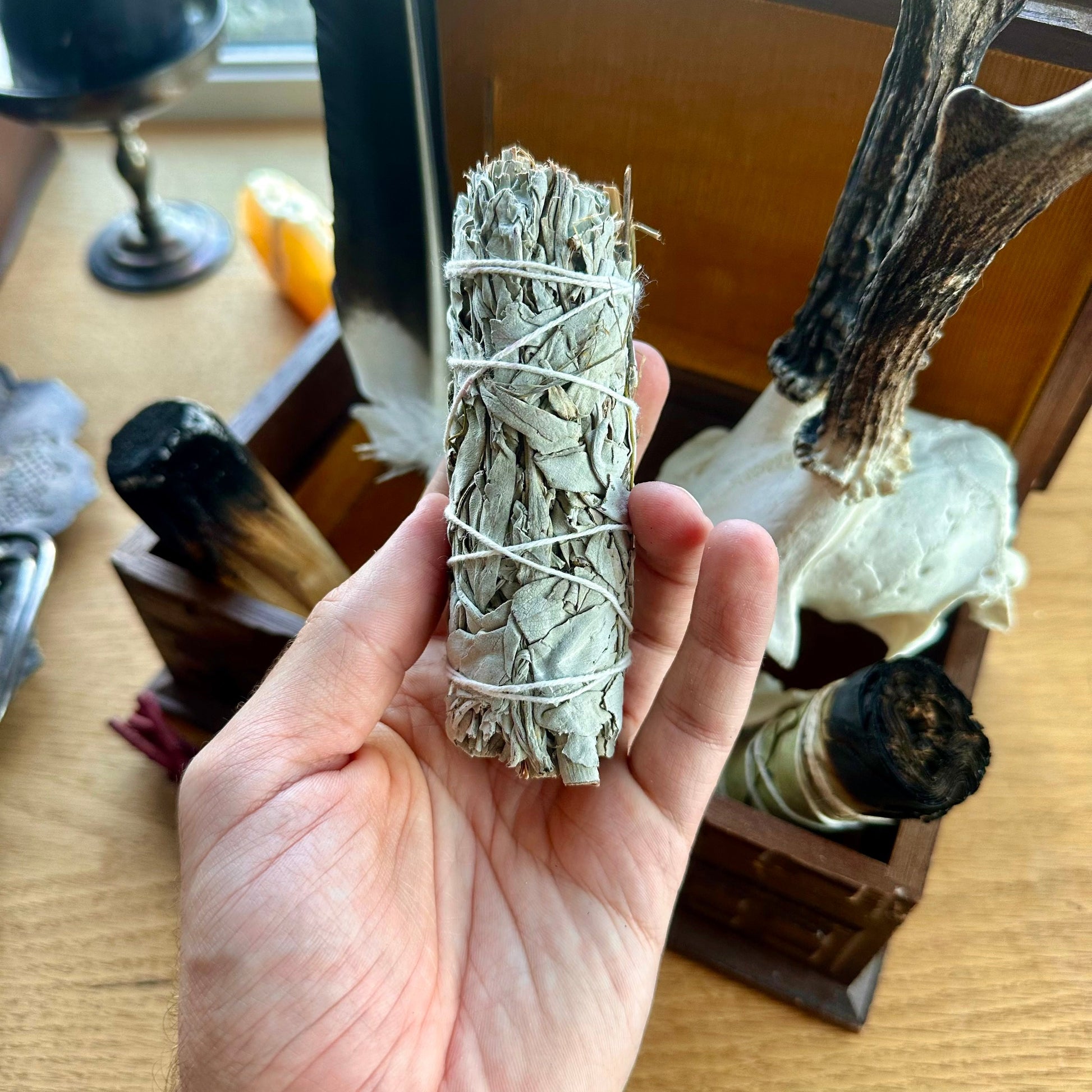 Hand holding a bundle of white sage wrapped in twine with a wooden surface and decorative items in the background.